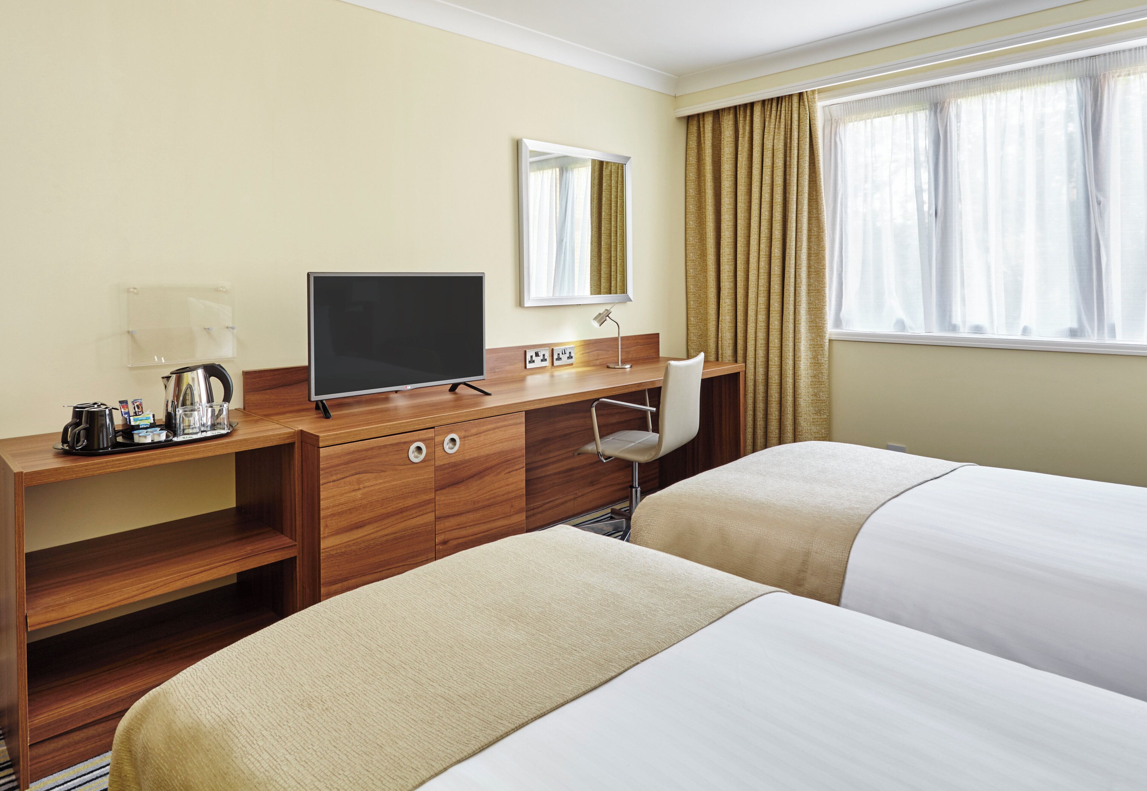 Een hotelkamer met een houten bureau, een flatscreen televisie, een koffiezetapparaat, twee bedden, een stoel en een raam met gordijnen.