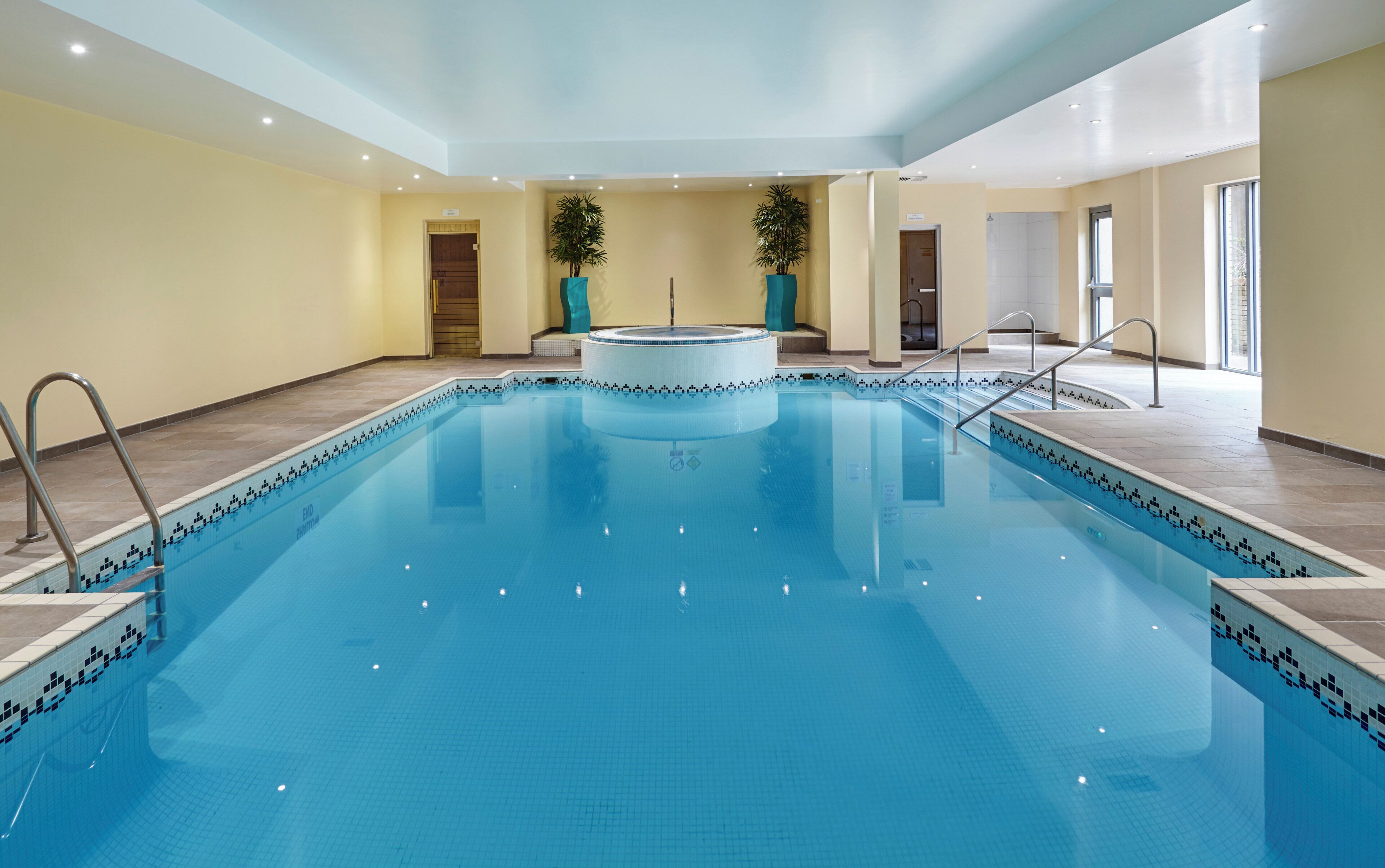 Ein großes Hallenbad mit gefliestem Rand, einer zentralen Whirlwanne und freiem Blick auf die umliegenden Wände und die Decke.