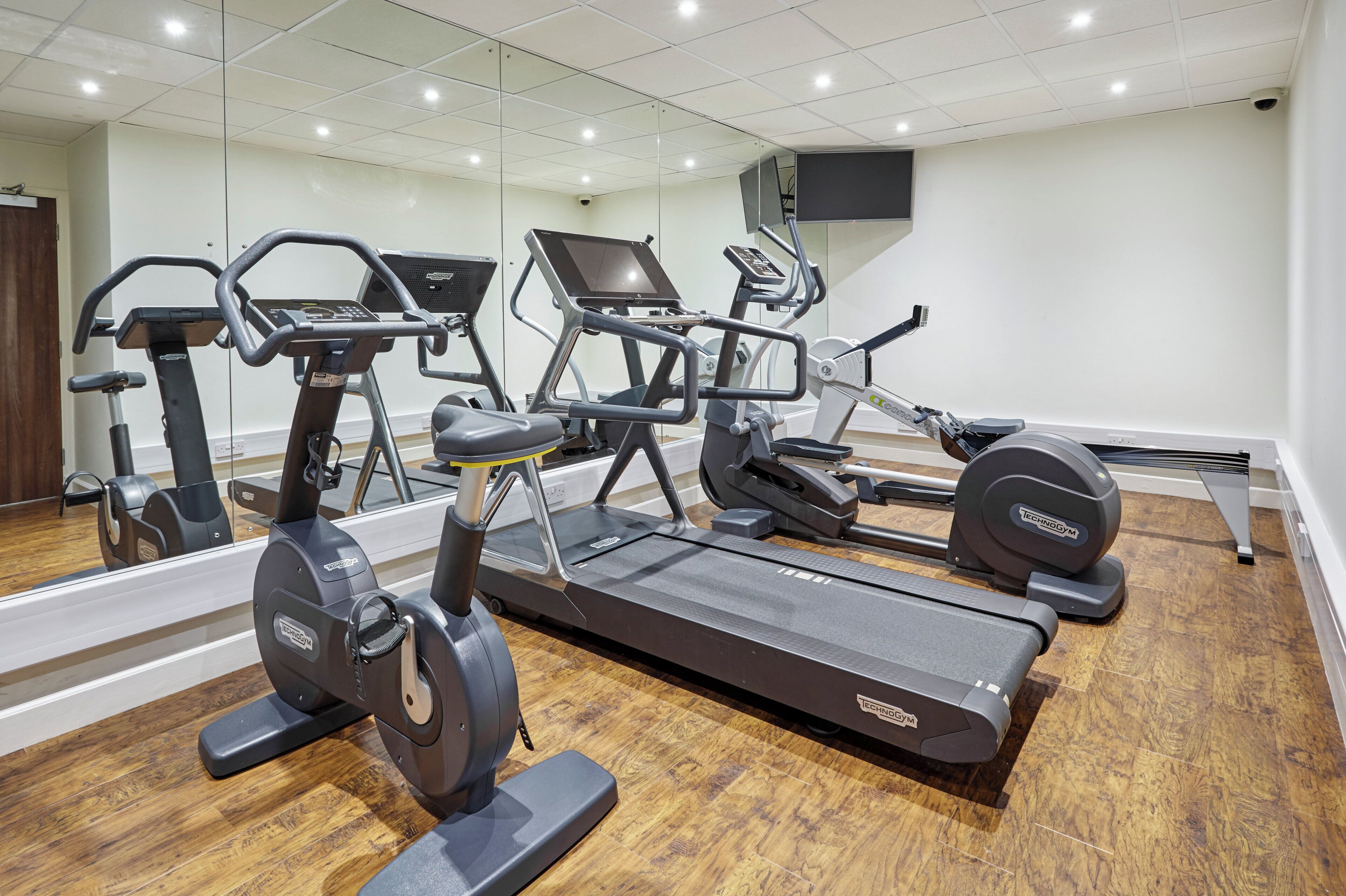 Ein Fitnessstudio mit mehreren Trainingsgeräten, einem Holzboden und einem großen Spiegel, der den Raum widerspiegelt.