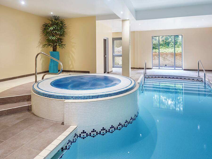 Uma grande piscina interior com hidromassagem, rodeada por paredes bege e uma planta em vaso.