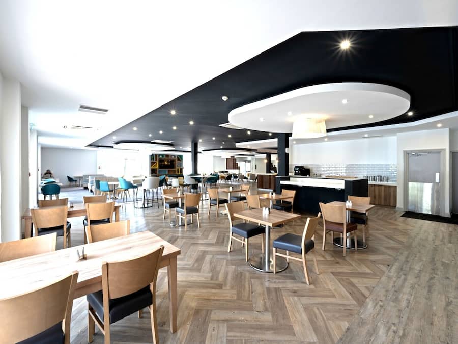 A modern dining area with wooden tables and chairs, a kitchen area with a counter, and a herringbone floor pattern.