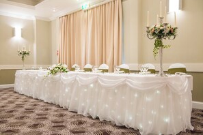 Un salón de banquetes con una larga mesa dispuesta para un evento formal, con manteles blancos, cristalería y centros de mesa florales.