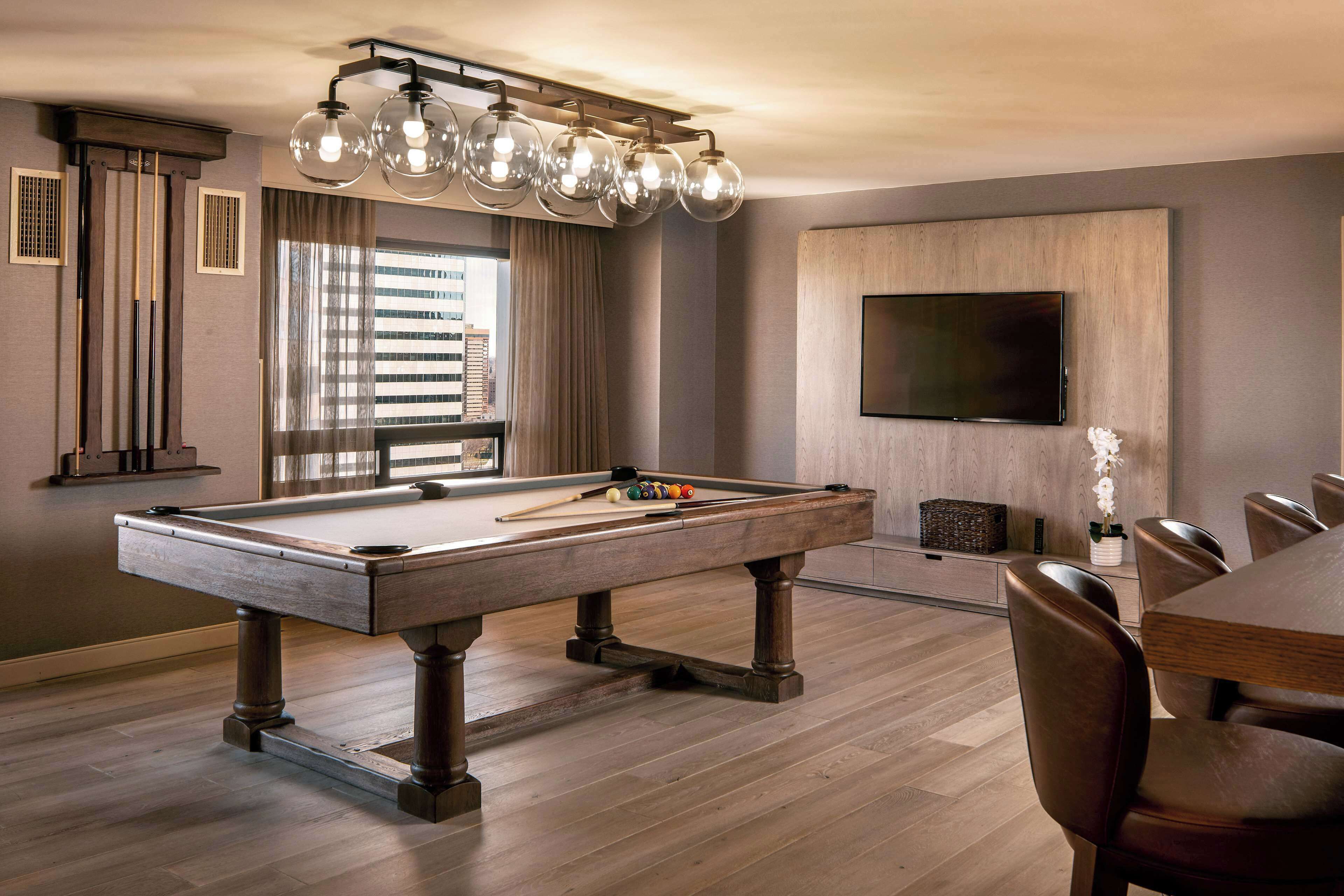 A pool table with a modern chandelier, a flat-screen TV mounted on the wall, and a dining area with leather chairs.