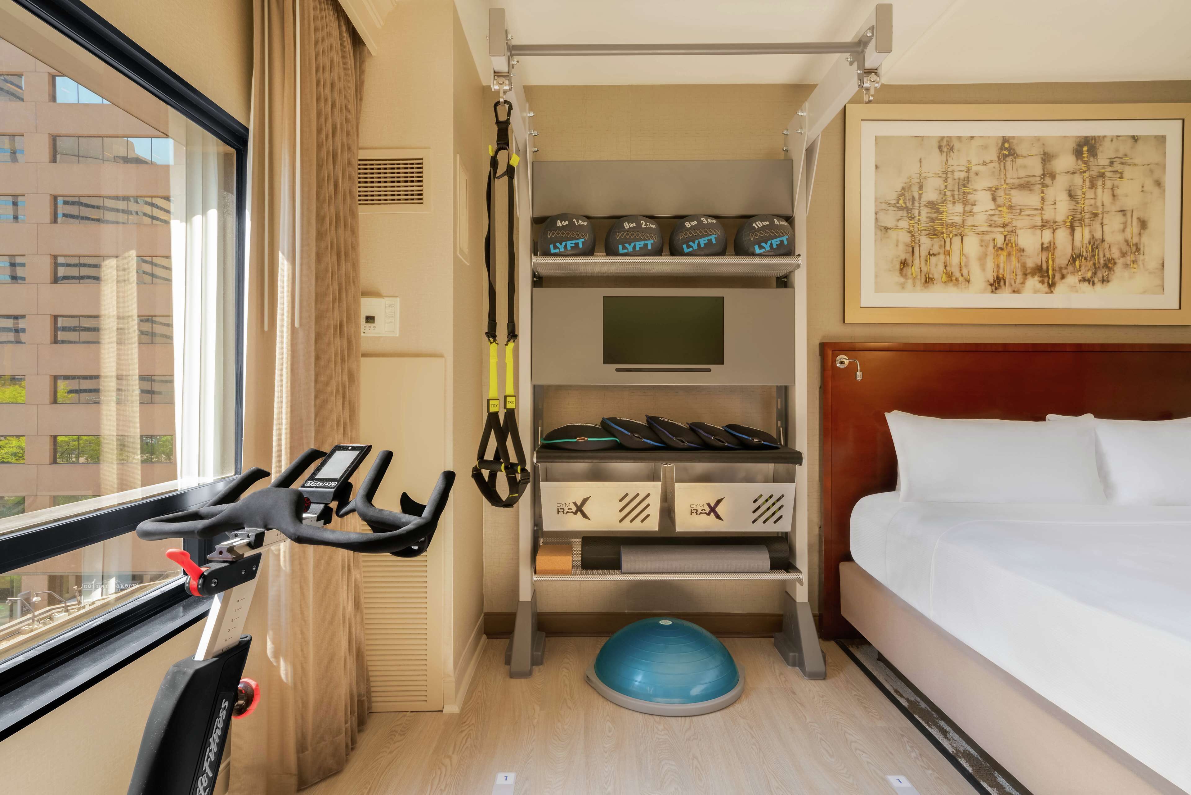 A hotel room with a bed, exercise equipment, a treadmill, a rowing machine, and a storage unit with exercise balls.