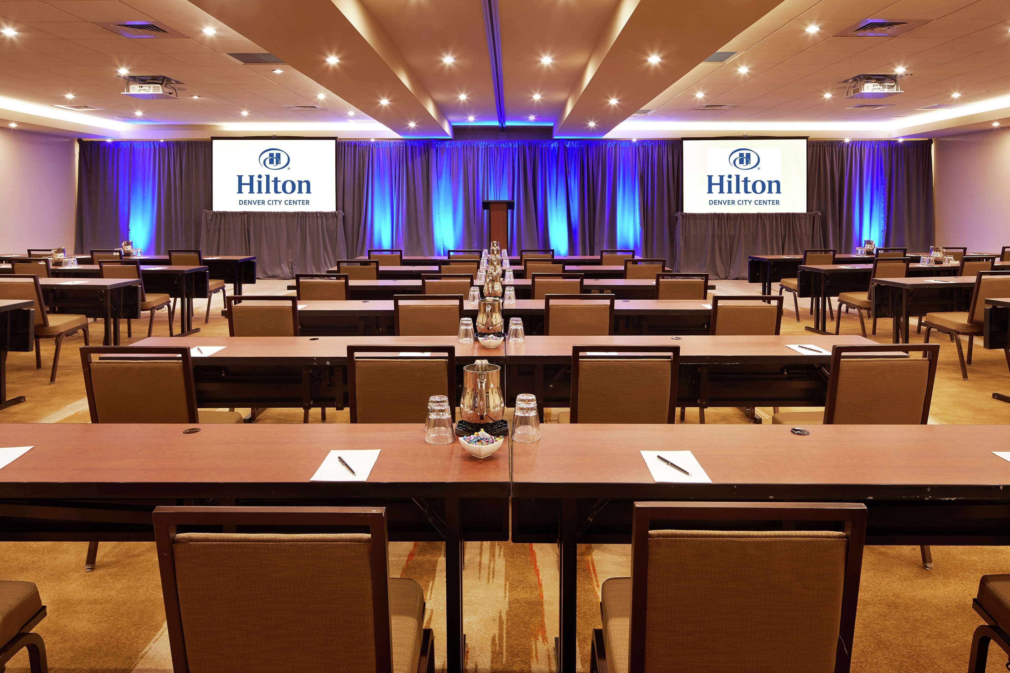 Una sala de conferencias en el Hilton Denver City Center con varias mesas y sillas, un atril y una pantalla grande que muestra el nombre del hotel.