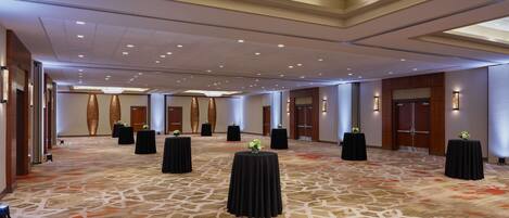A spacious hotel lobby with a patterned carpet, multiple black tables with floral centerpieces, and a high ceiling with recessed lighting.