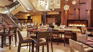 Um interior de restaurante moderno com uma lareira central, várias mesas de jantar e uma escadaria que leva a um andar superior.