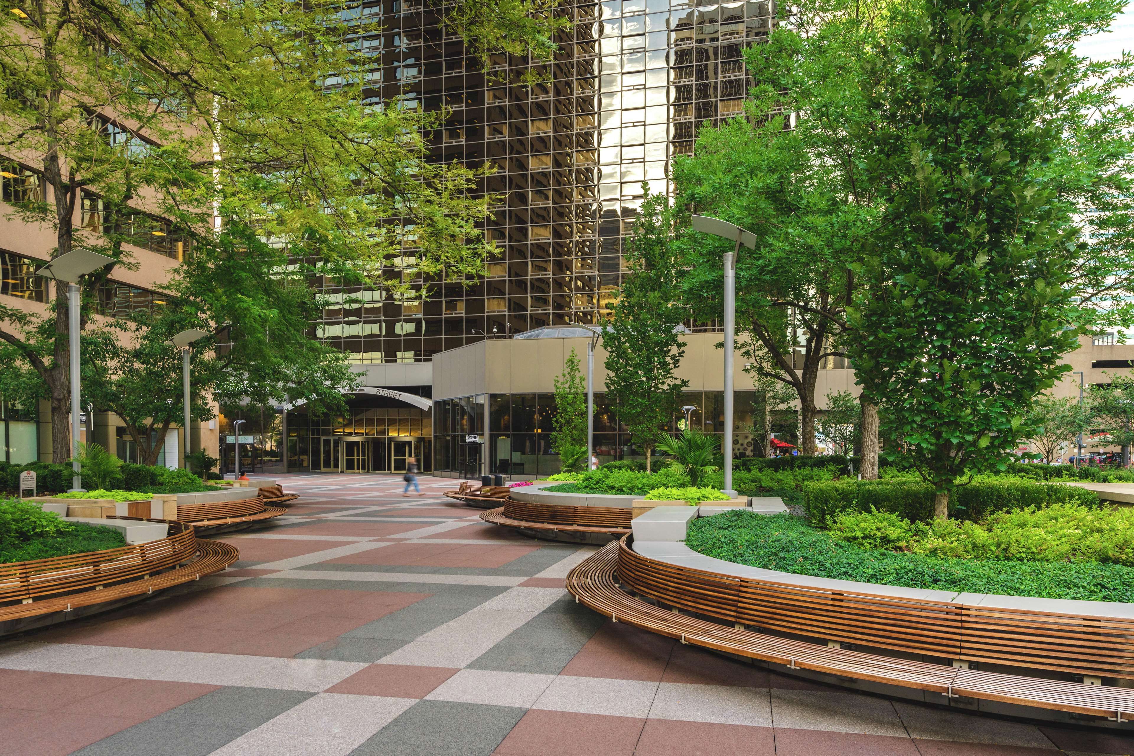 A modern urban plaza with a central seating area, surrounded by greenery and a tall building in the background.