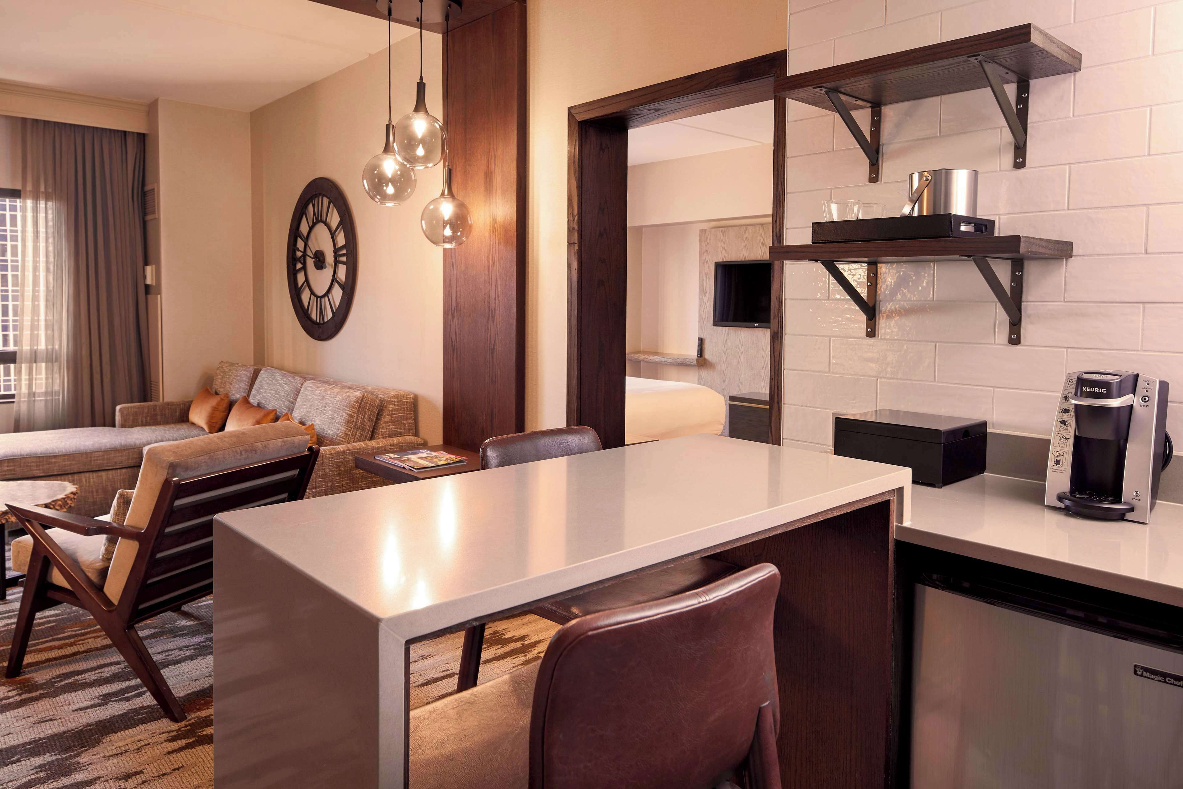 A modern hotel room with a dining area, a sofa, a coffee machine, and a TV mounted on the wall.