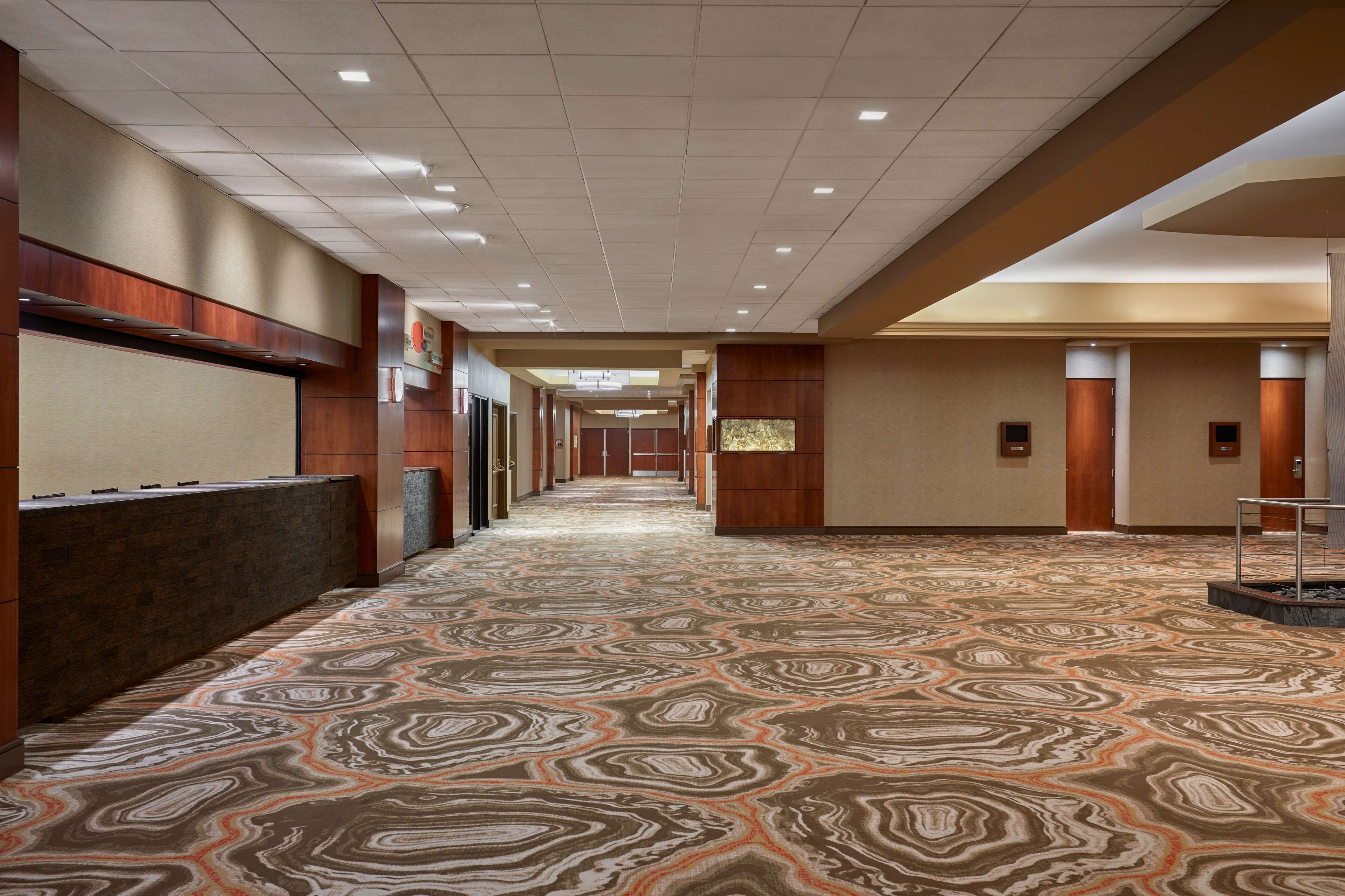A hotel corridor with patterned carpet, wooden paneling, and recessed lighting.