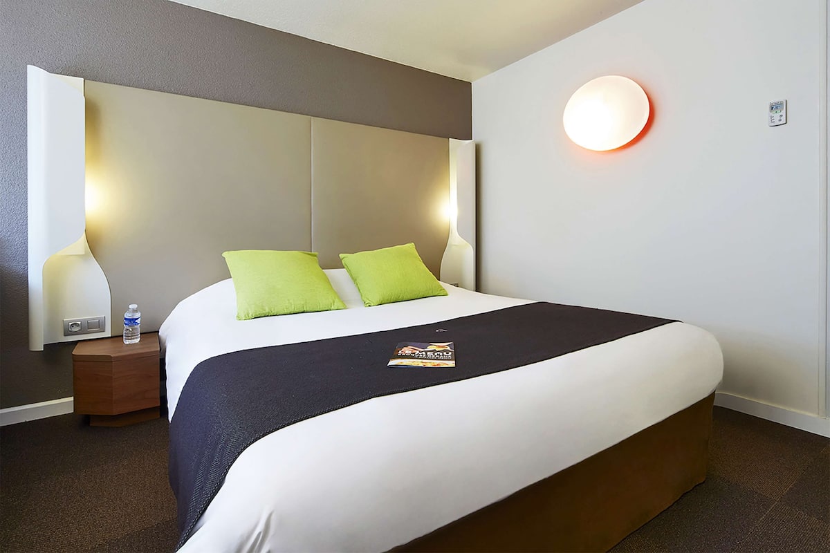 A neatly made bed with a dark headboard, two green pillows, and a black blanket. There is a small wooden nightstand with a water bottle and a lamp beside the bed.