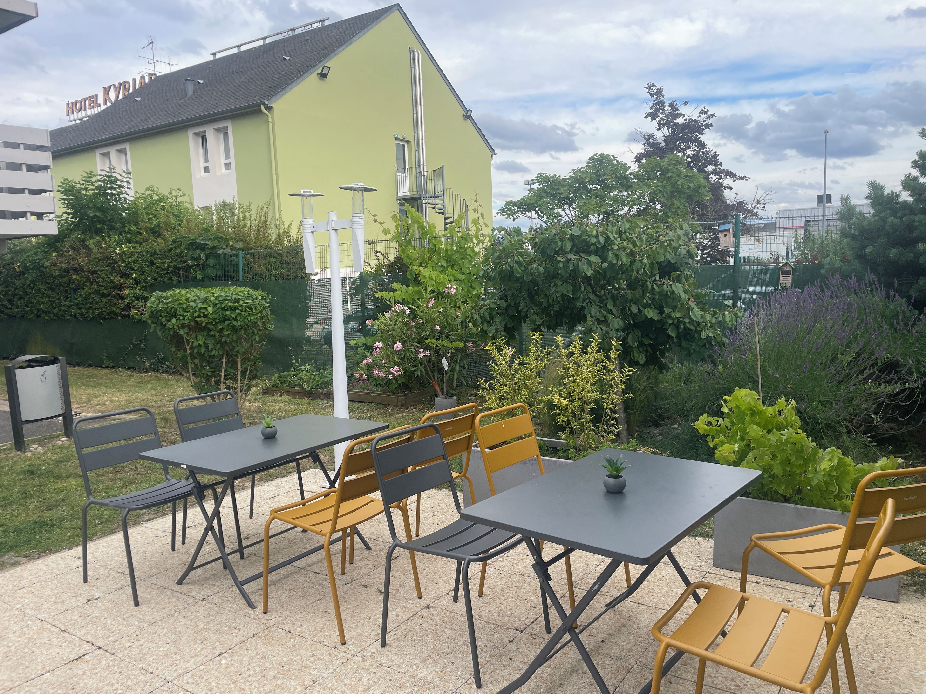 An outdoor seating area with tables and chairs, a green building in the background, and a variety of plants.