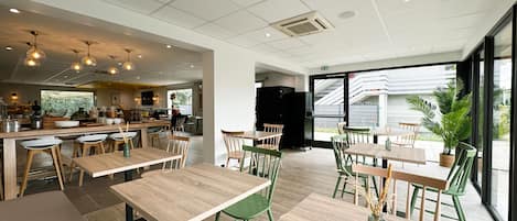 A modern cafe interior with wooden tables and chairs, a kitchen area, and large windows offering a view of the outside.