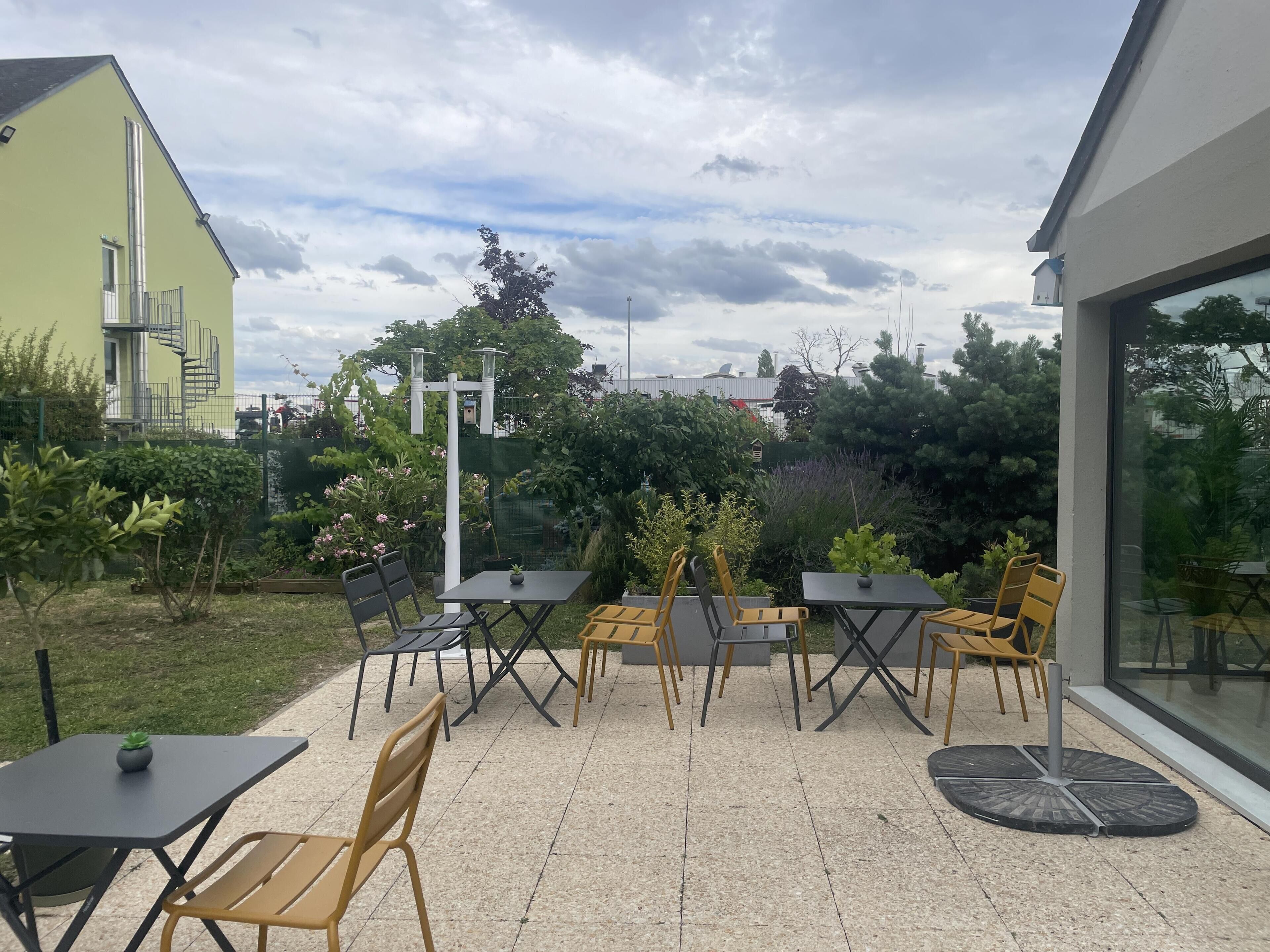 Une terrasse extérieure avec des tables et des chaises, entourée de verdure et bordée d’un bâtiment dont on peut voir une fenêtre.