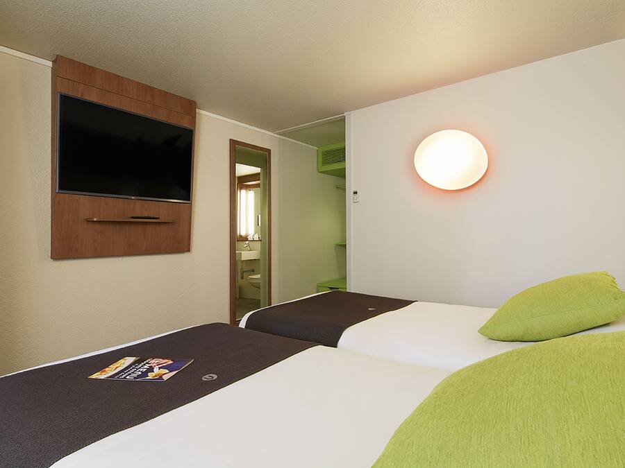 A hotel room with two beds, a TV mounted on the wall, a bedside table with a magazine, and a round wall light.