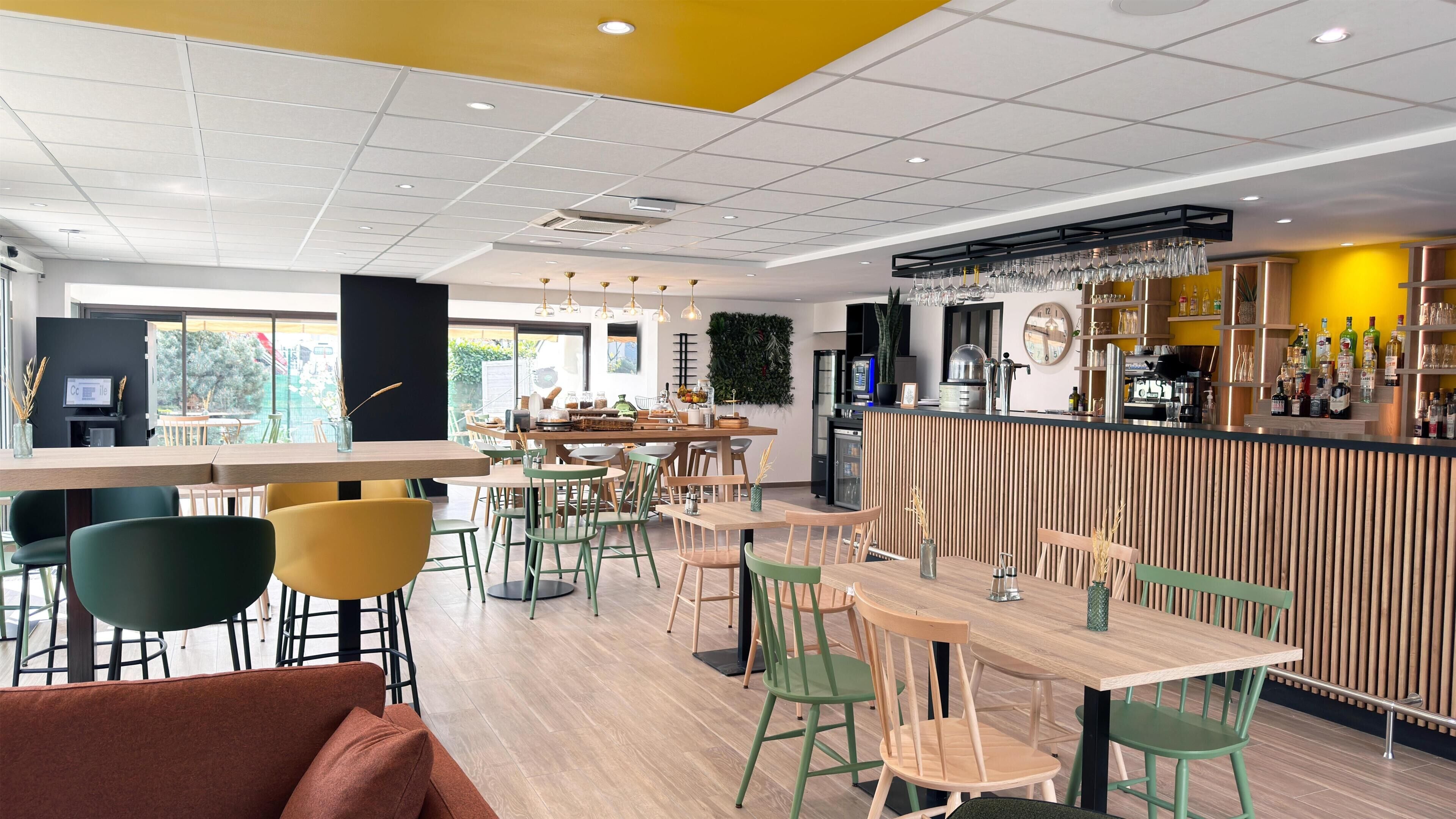 Een modern restaurant met een bar, houten meubels en een groene wand met planten.