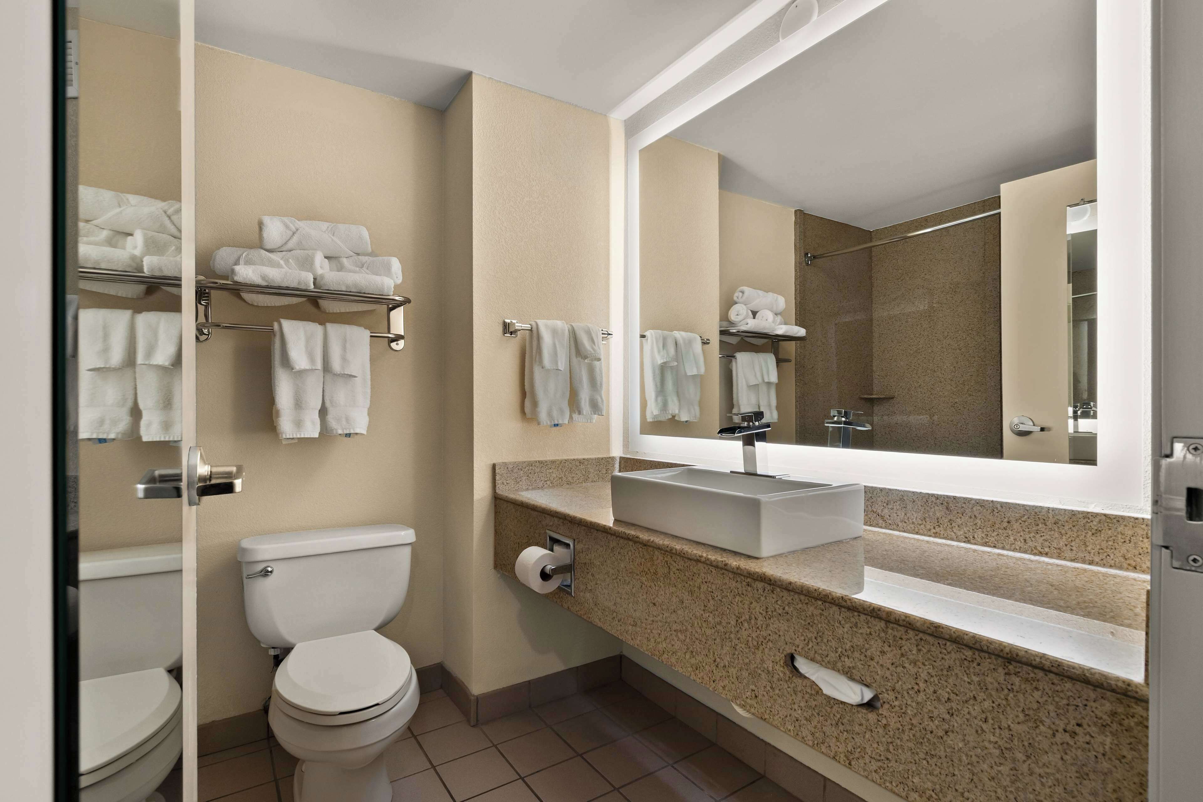 A modern bathroom with a toilet, sink, and mirror. There are towels on a rack and a shower area visible in the reflection.