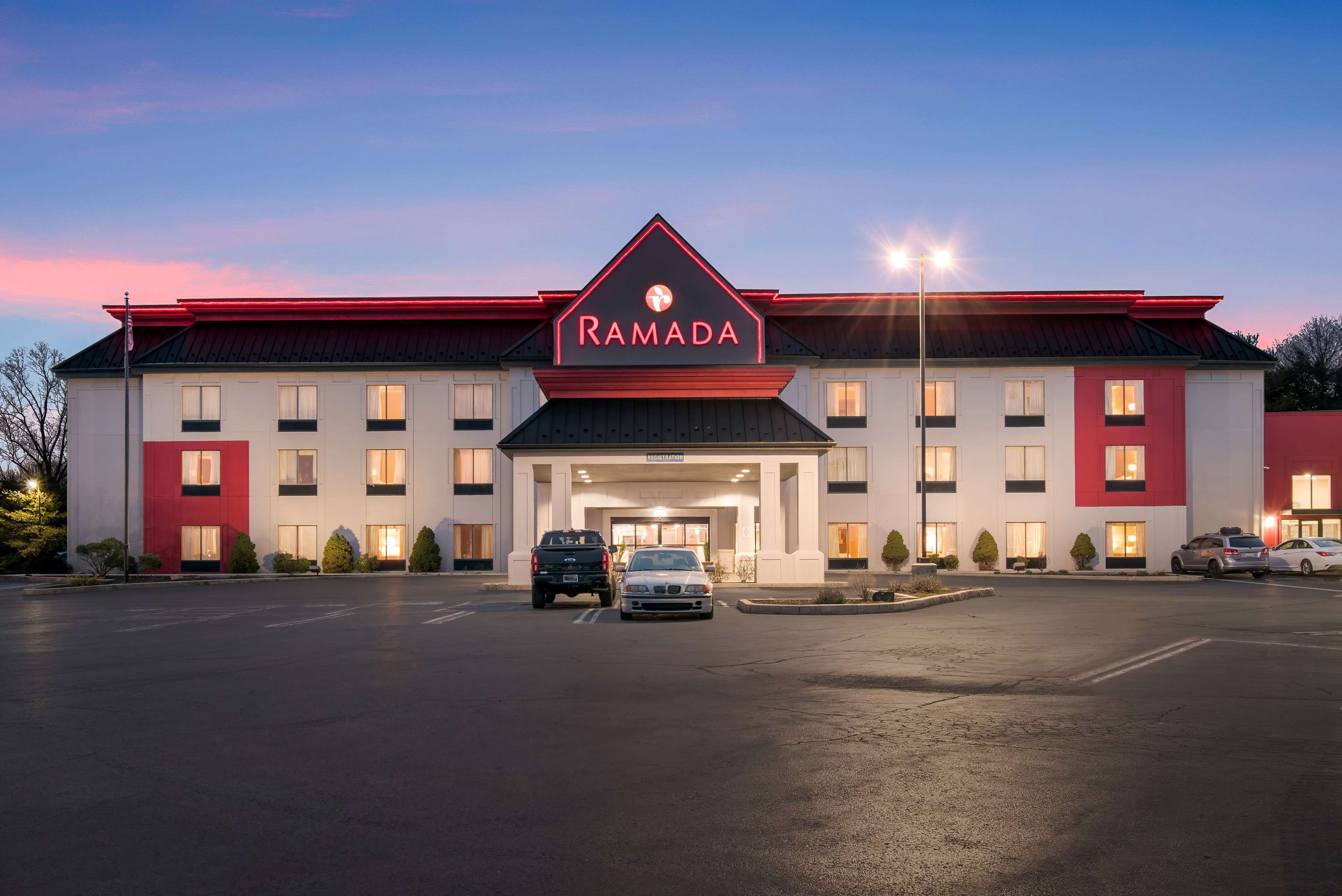 Un hôtel à plusieurs étages, reconnaissable à son grand panneau « RAMADA », avec un parking et plusieurs véhicules stationnés devant.