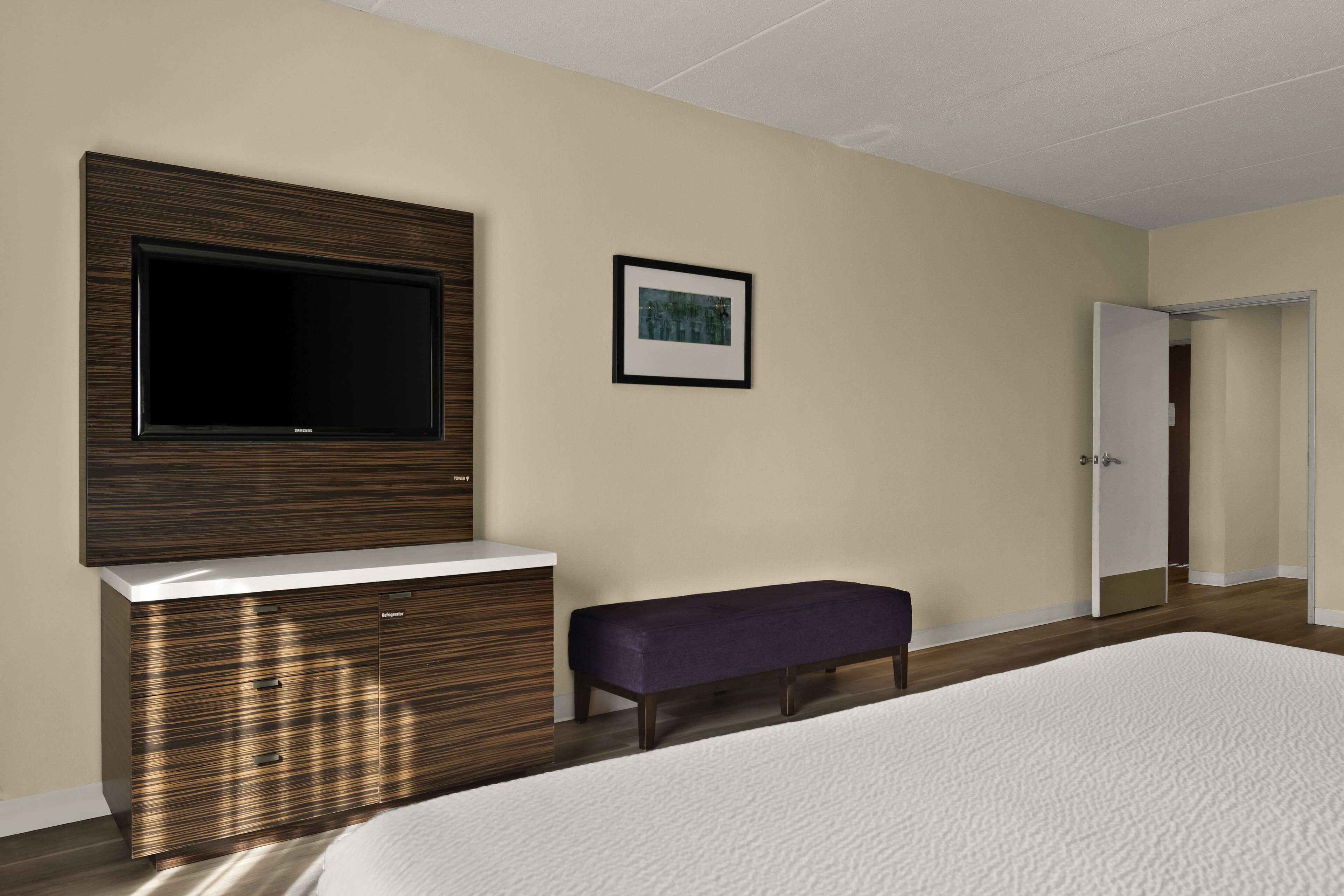 A bedroom with a bed, a TV mounted on a wooden cabinet, a bench, and a door leading to another room.