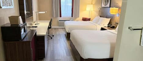 Ein Hotelzimmer mit zwei Betten, einem Schreibtisch, einem Fernseher und einem Fenster mit Vorhängen.
