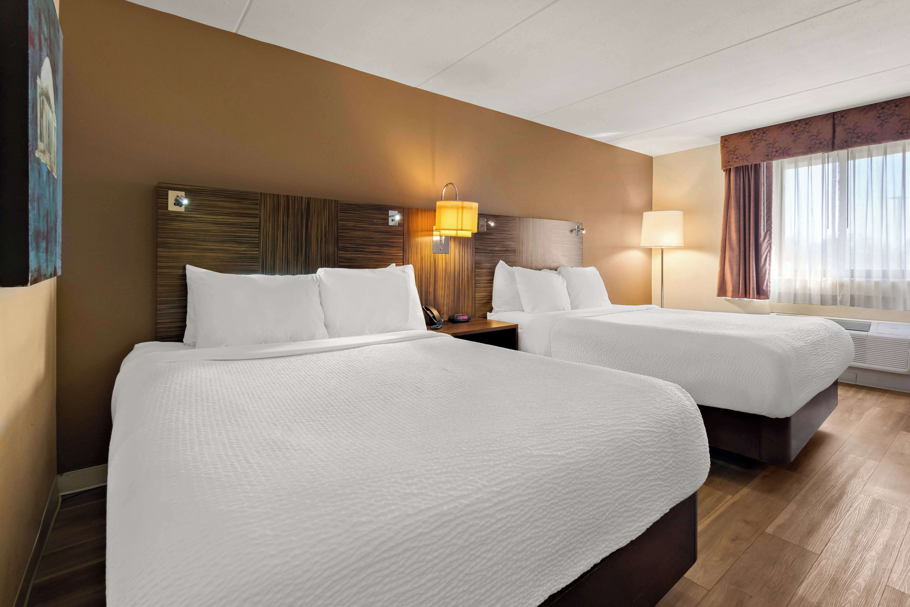 A hotel room with two beds, a wooden headboard, a nightstand, a lamp, and a window with curtains.