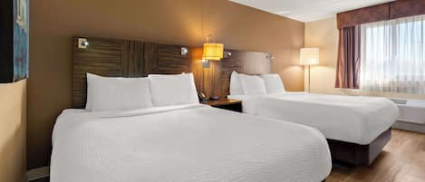 A hotel room with two beds, a wooden headboard, a nightstand, a lamp, and a window with curtains.