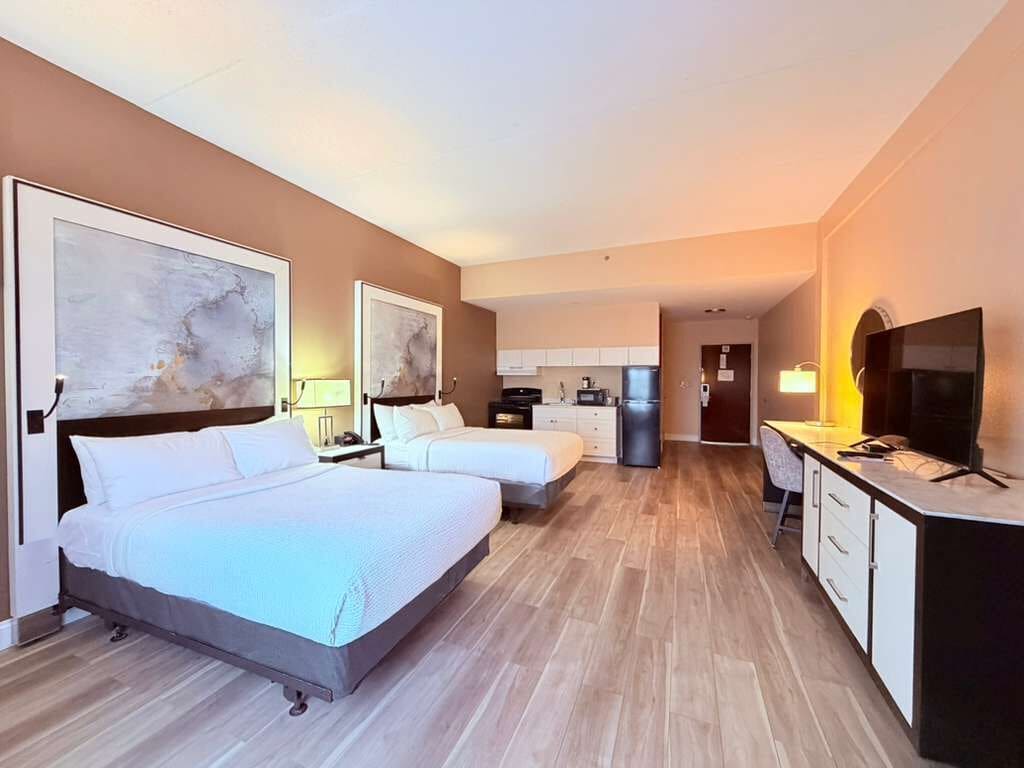 A hotel room with two beds, a desk, a TV, and a kitchenette.