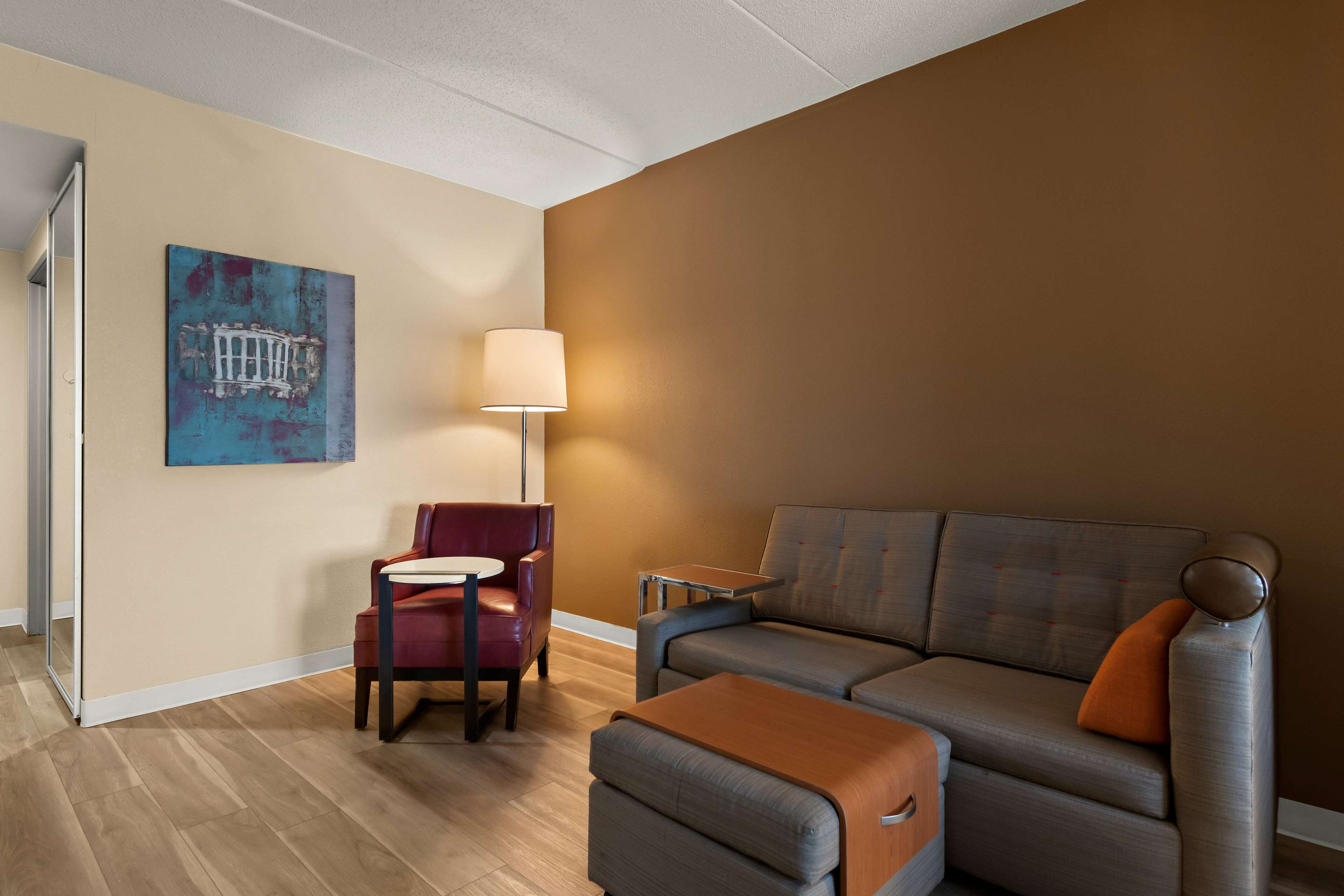 A modern hotel room with a sofa, armchair, and ottoman, a small table, and a lamp.