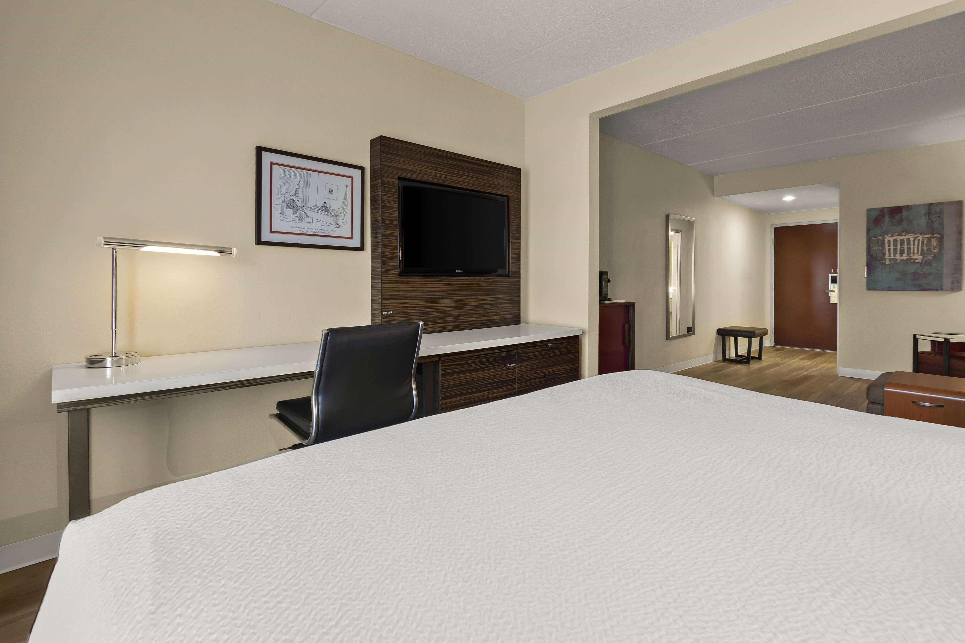 A hotel room with a large bed, a desk with a lamp, a flat-screen TV mounted on the wall, and a small table.
