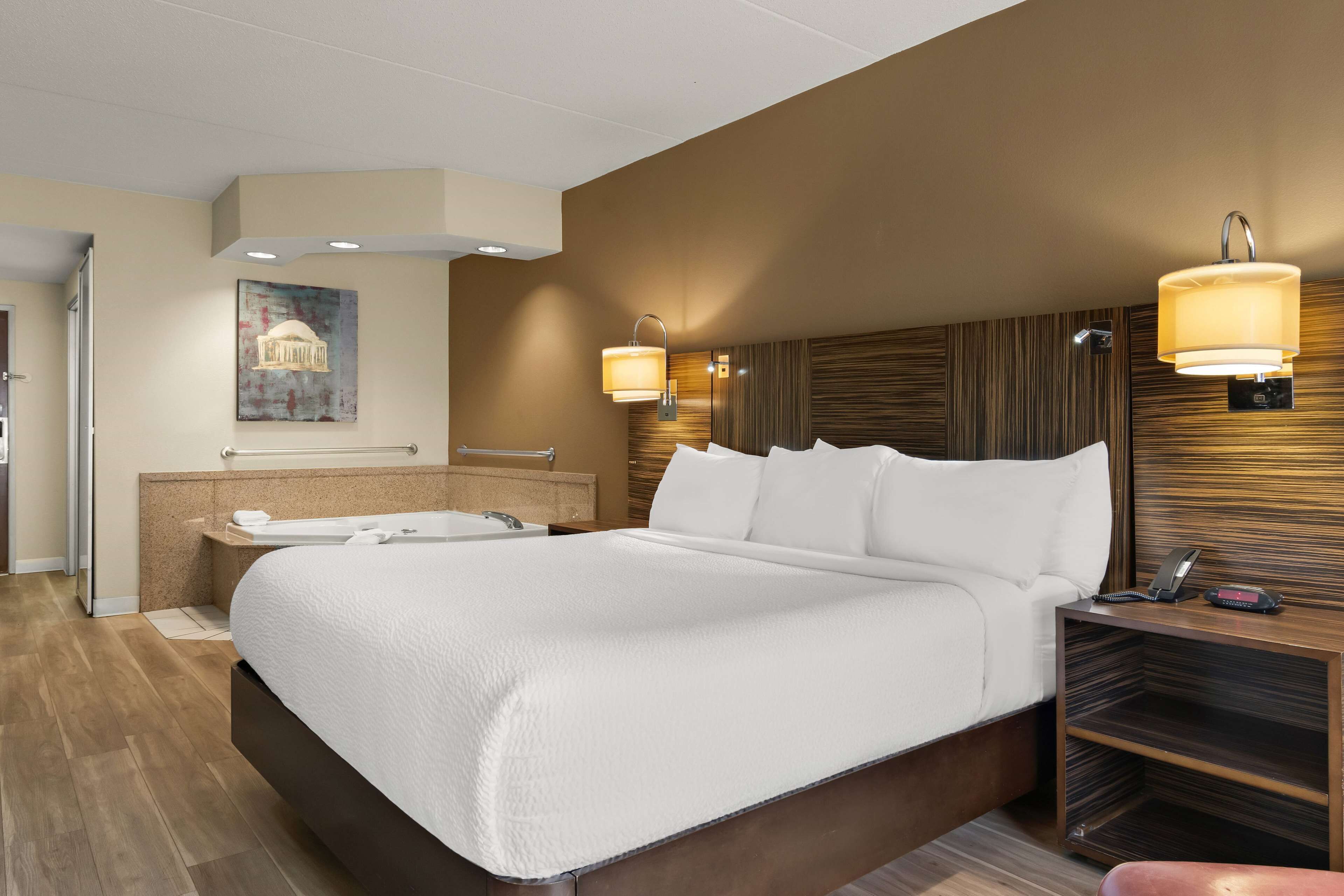 A hotel room with a large bed, a built-in bathtub, a vanity with a mirror, and two wall-mounted lamps.