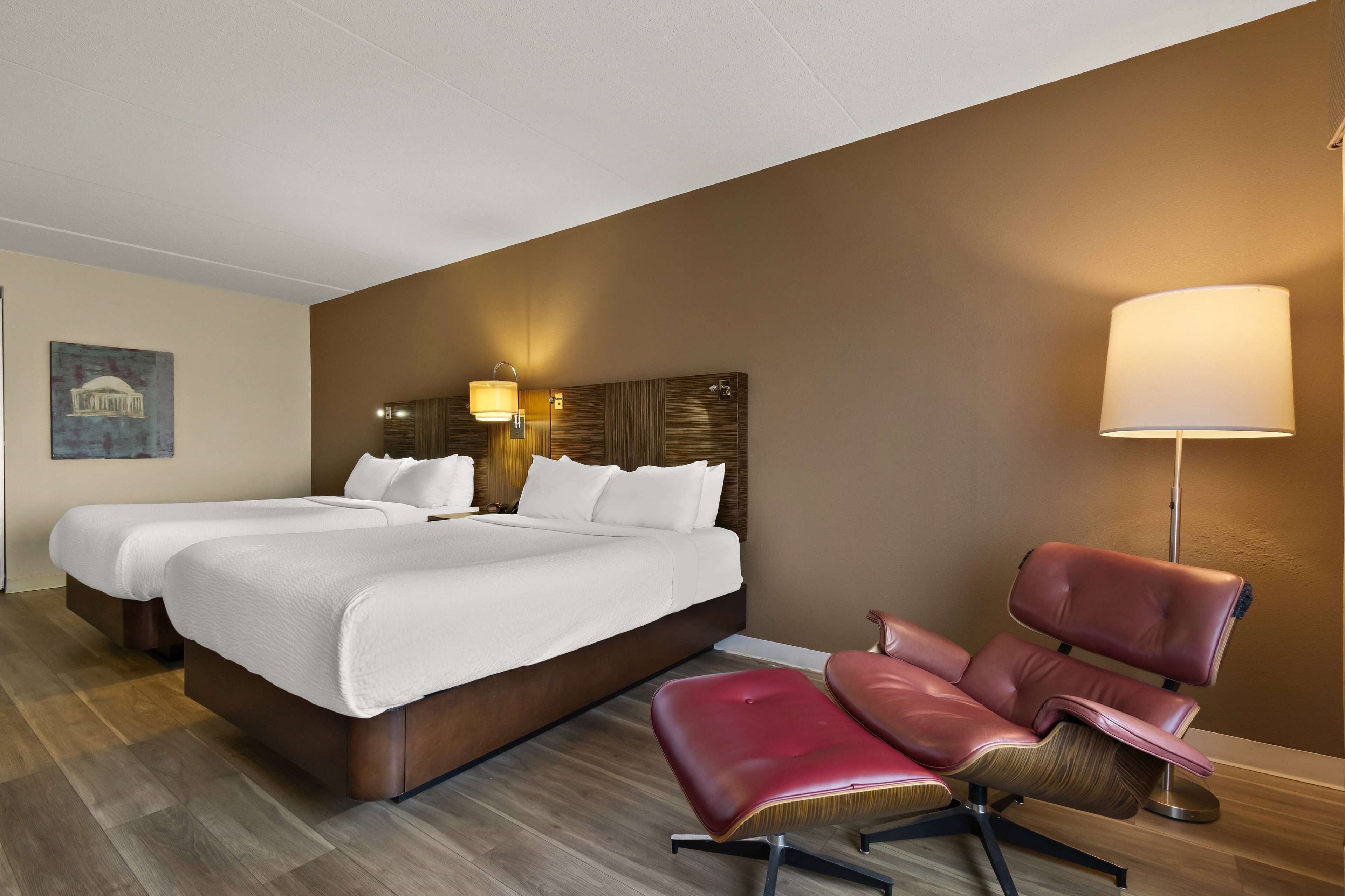 Una habitación de hotel con dos camas, un sillón de cuero rojo, una lámpara de pie y un cuadro en la pared.