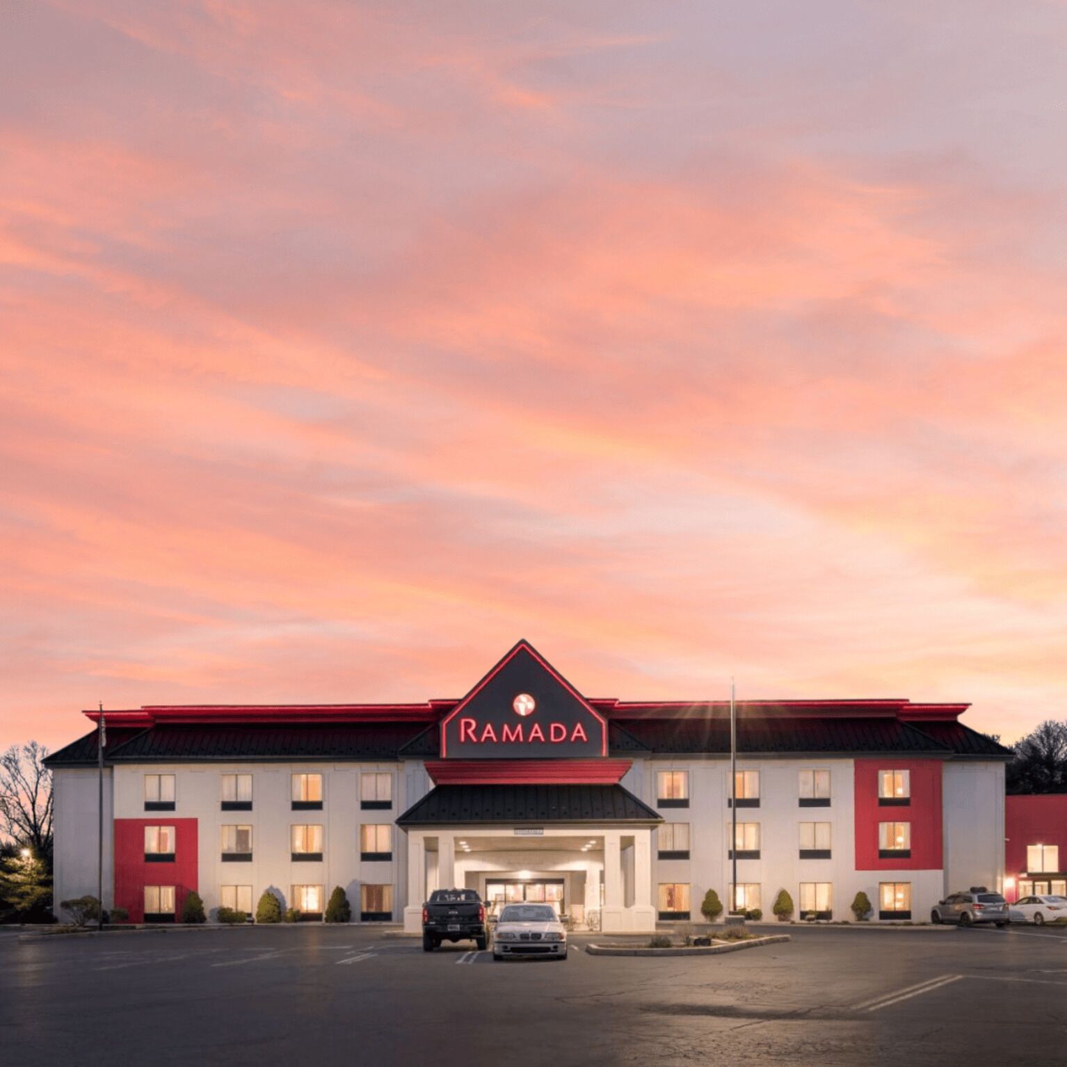 A Ramada hotel with a modern design, featuring a prominent red and white color scheme, a large parking lot, and a clear sky.