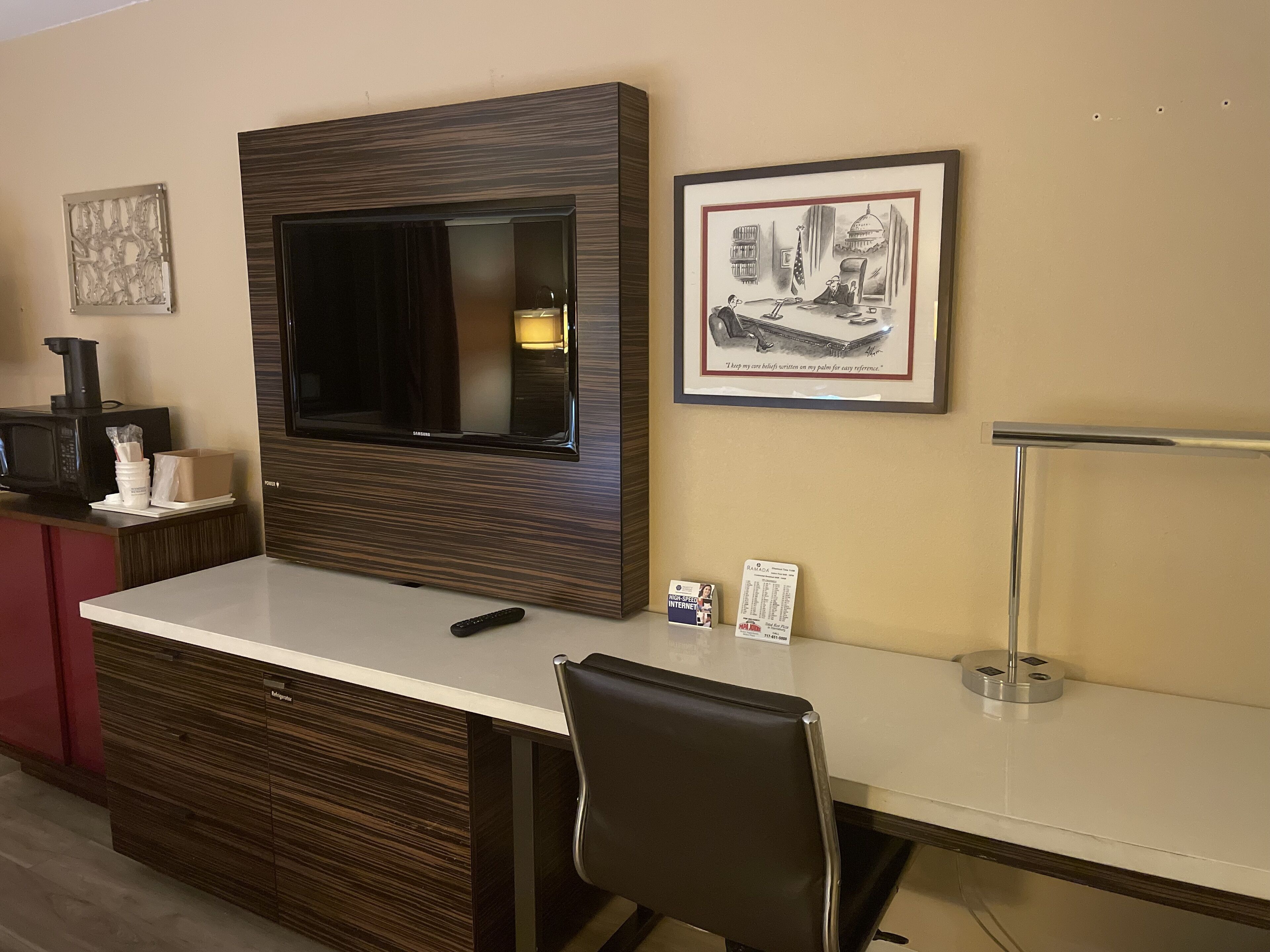 A hotel room with a large flat-screen TV mounted on the wall, a desk with a chair, a microwave, and a coffee machine on the counter.