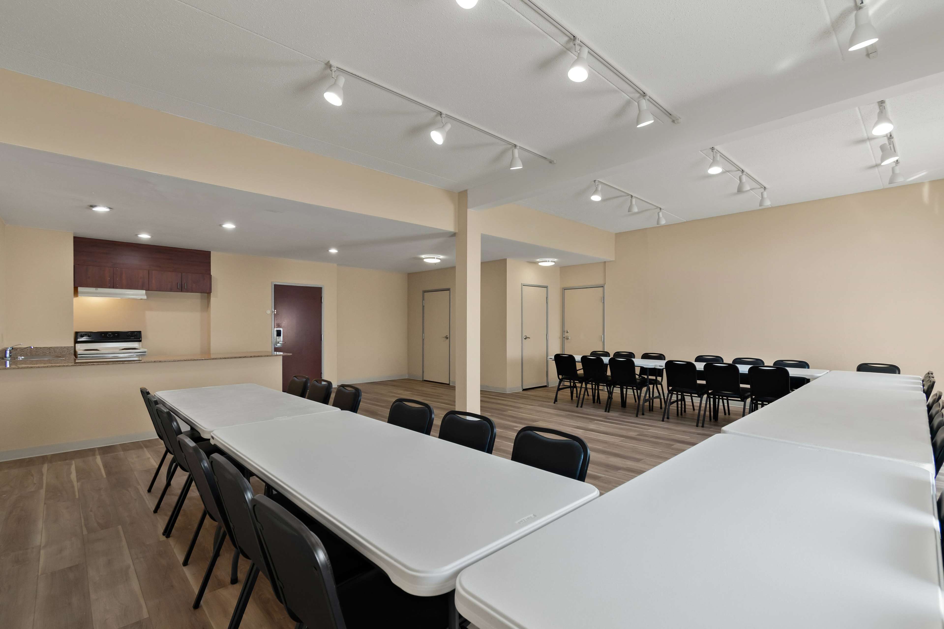 A conference room with long tables and chairs, recessed ceiling lights, and a kitchenette area.