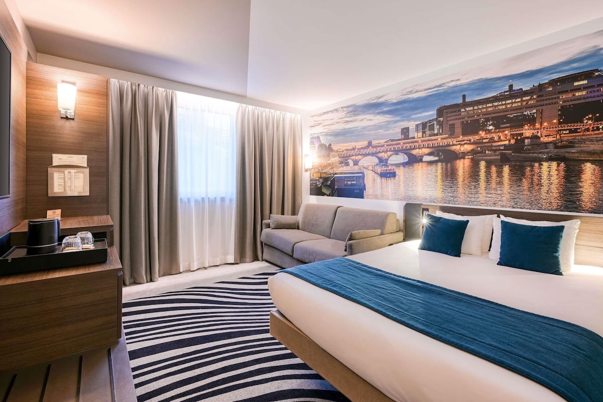 A hotel room with a bed, a sofa, a desk, and a view of a river at sunset.
