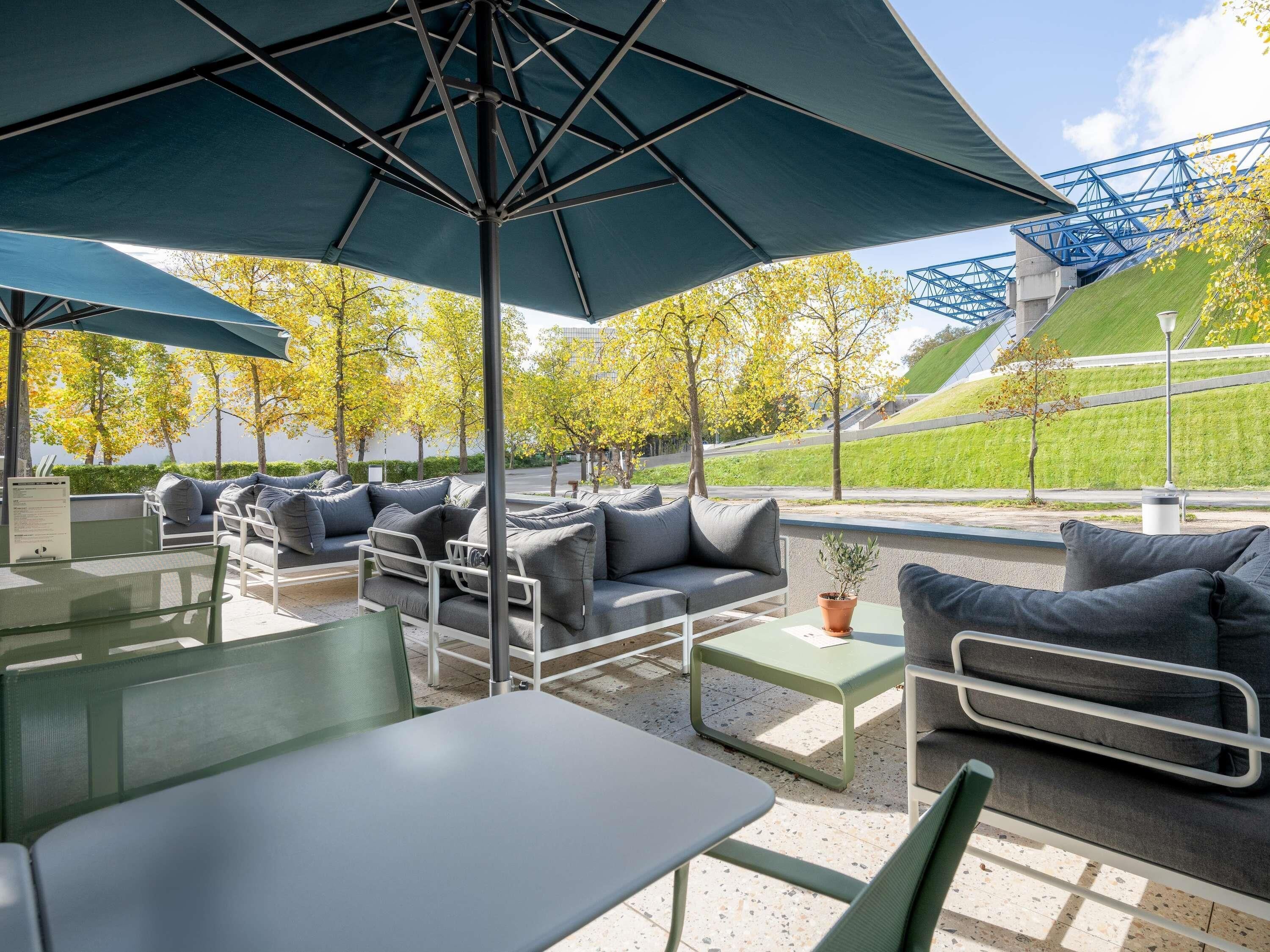 An outdoor seating area with tables and chairs, a large umbrella, and a view of a green hill and trees.