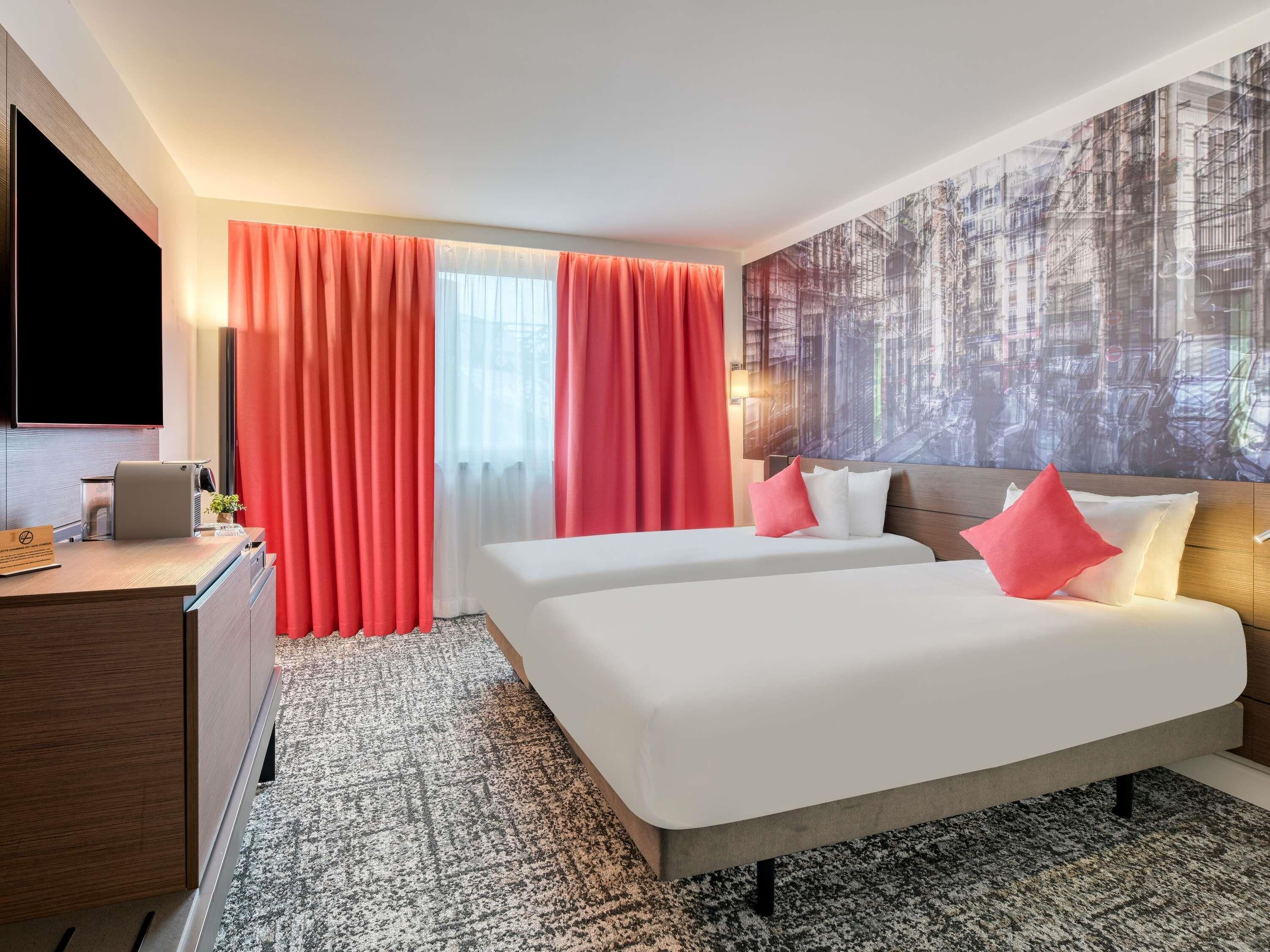 A hotel room with two beds, a desk, a TV, and a large window with curtains.