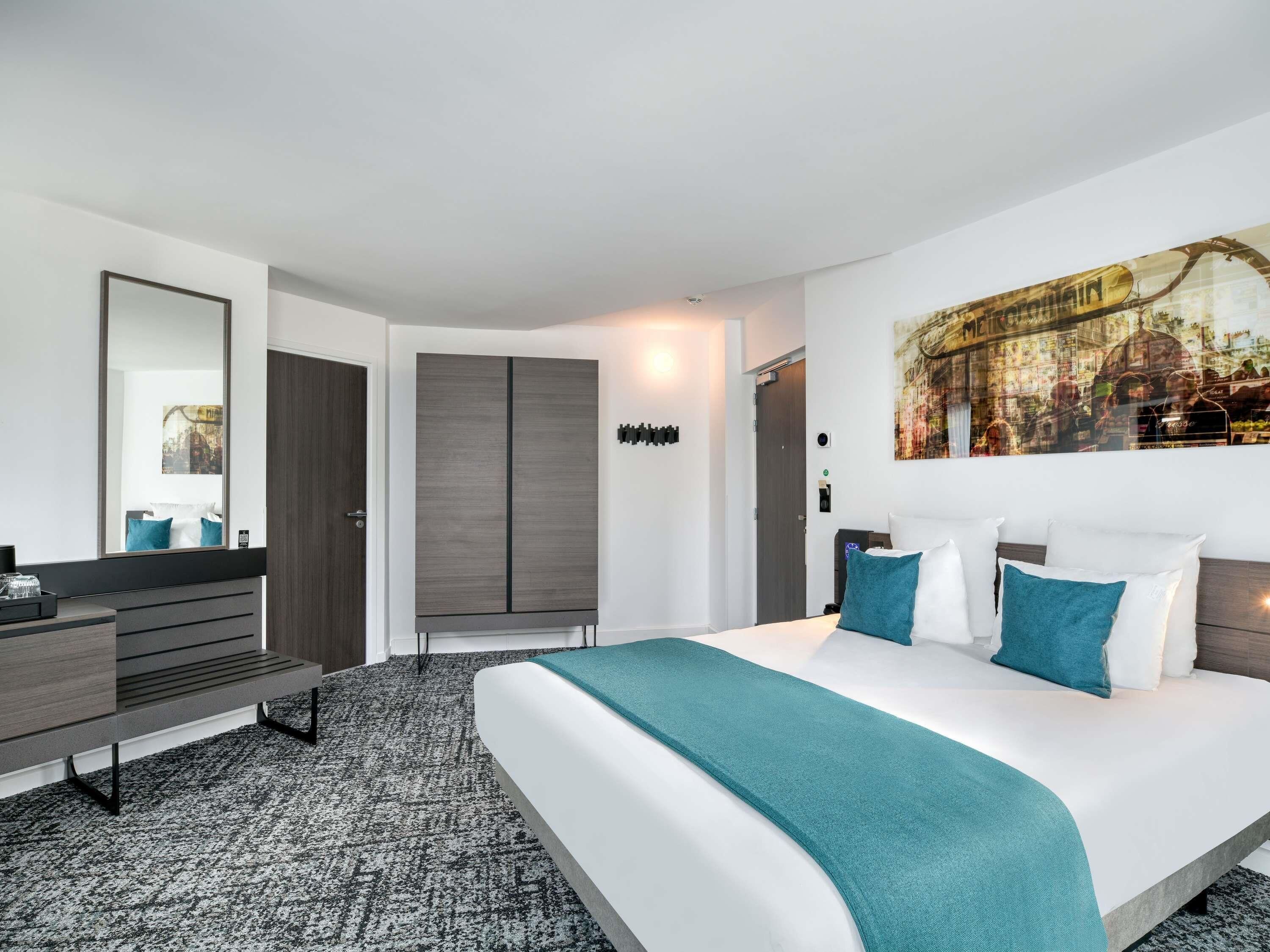 A modern hotel room with a large bed, a desk, and a wardrobe.