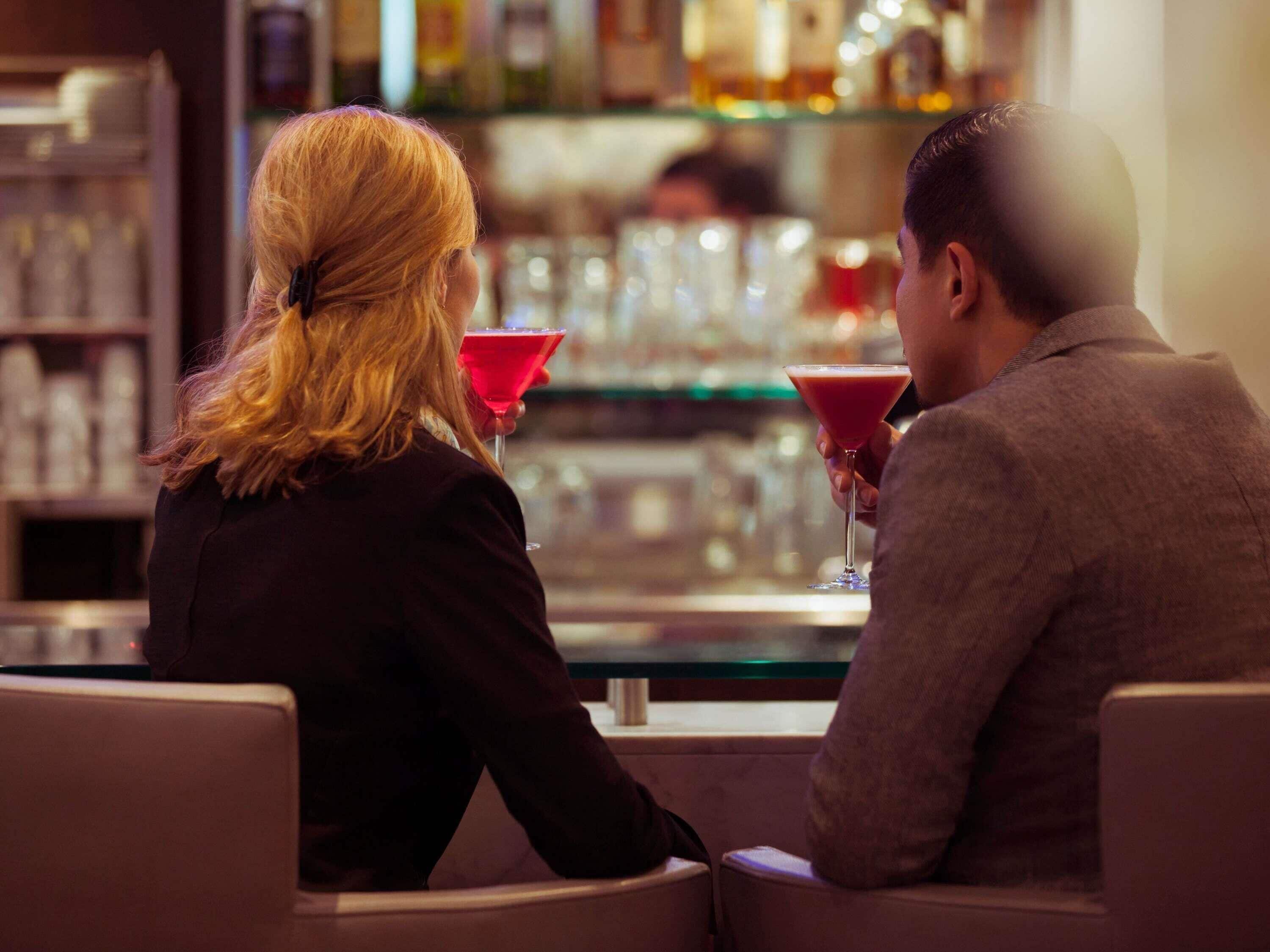 Two people seated at a bar with a glass of red drink in front of them.