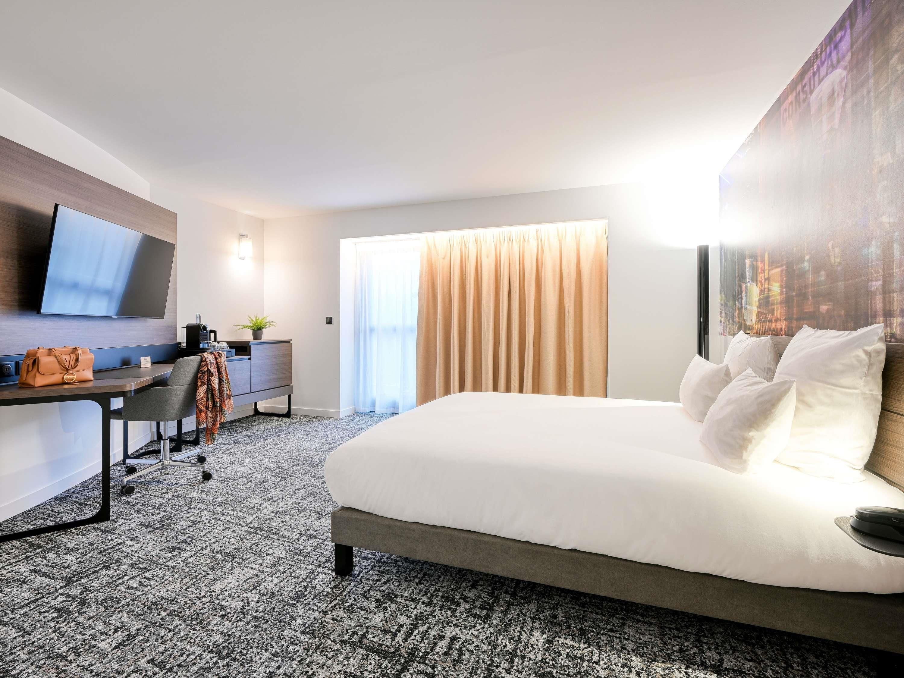 A hotel room with a bed, a desk with a chair, a flat-screen TV, a coffee machine, and a window with curtains.