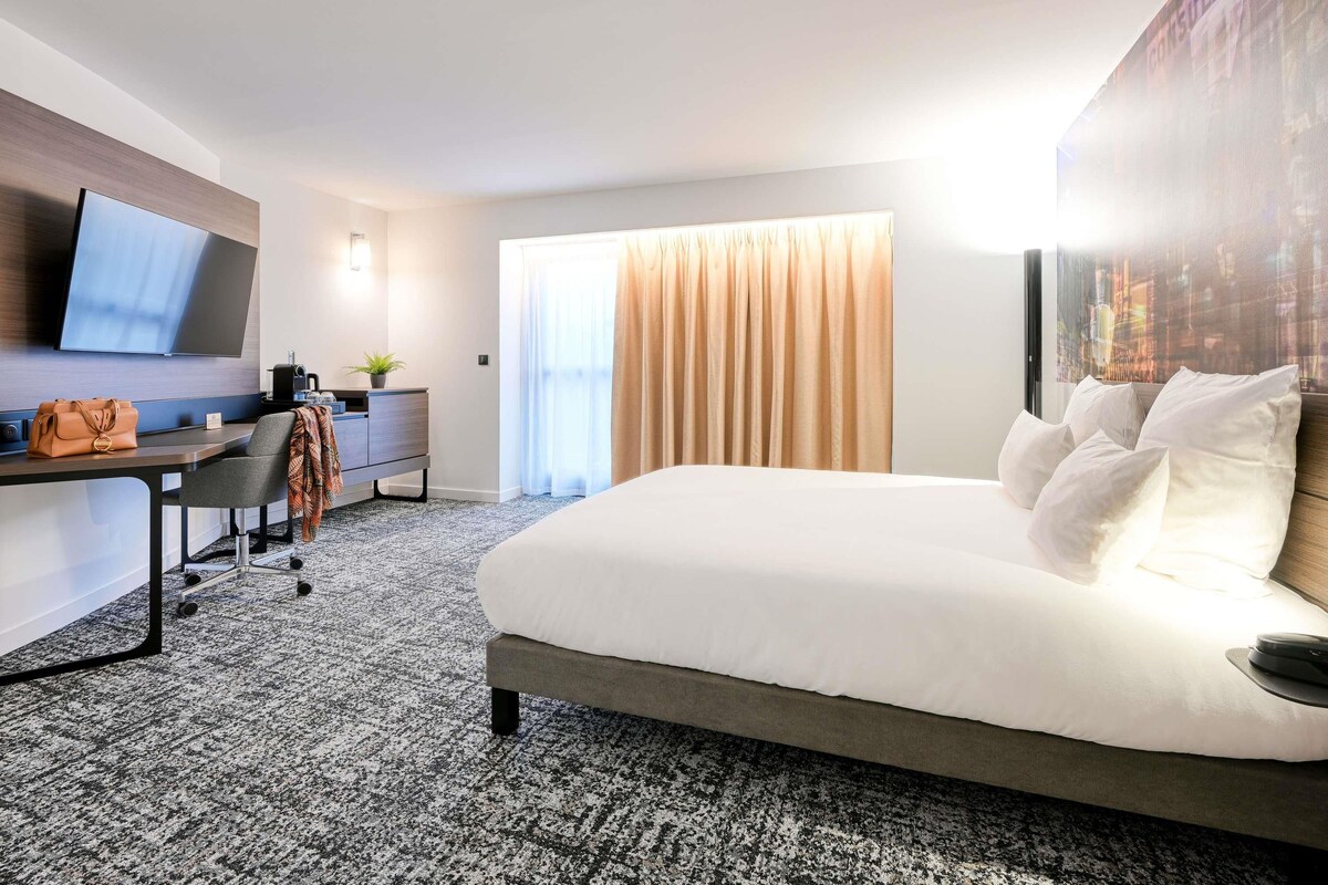 A hotel room with a bed, a desk with a chair, a flat-screen TV, a coffee machine, and a window with curtains.