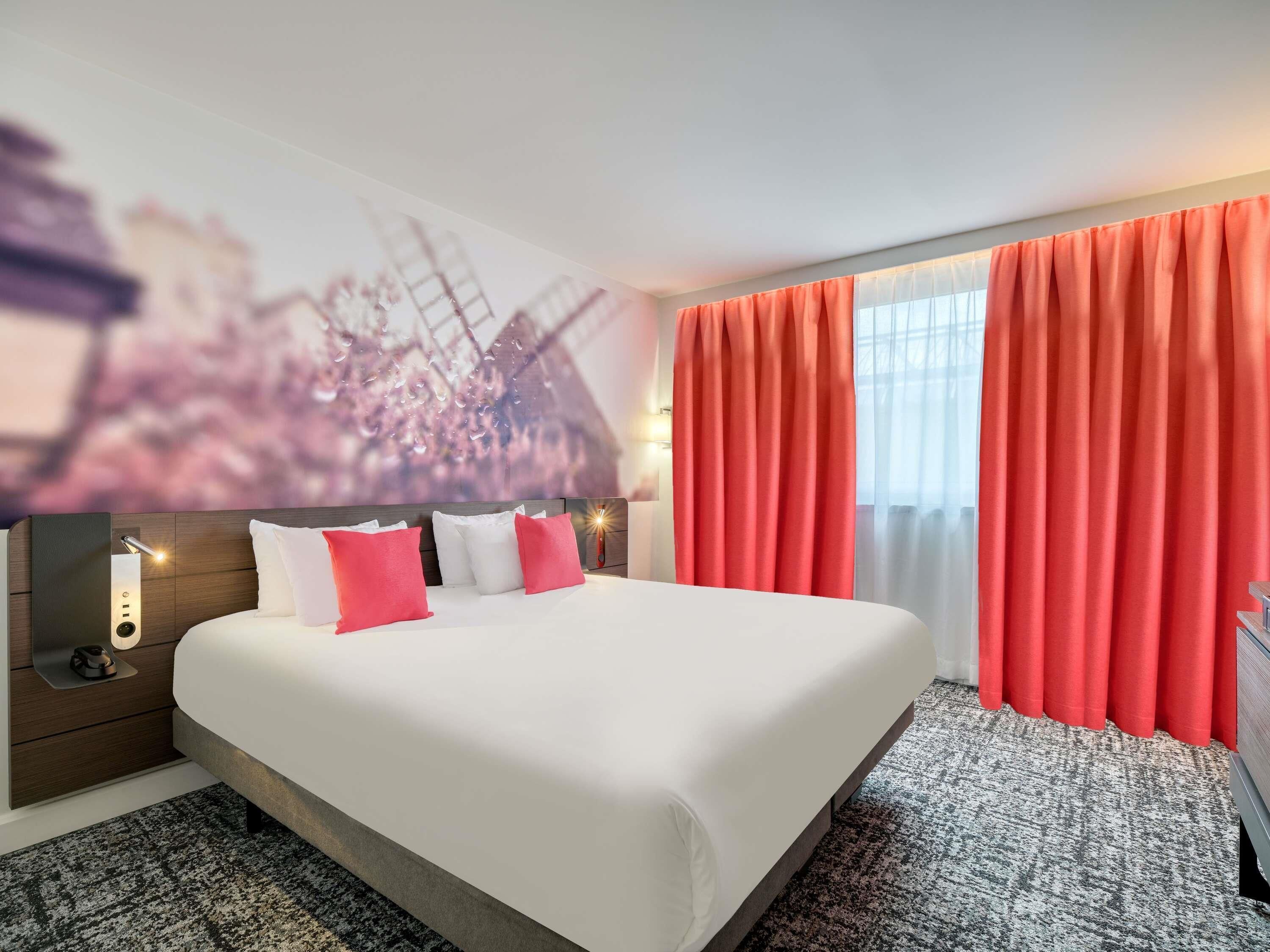 Une chambre d’hôtel avec un grand lit, des rideaux rouges, un téléviseur fixé au mur et une moquette à motifs.
