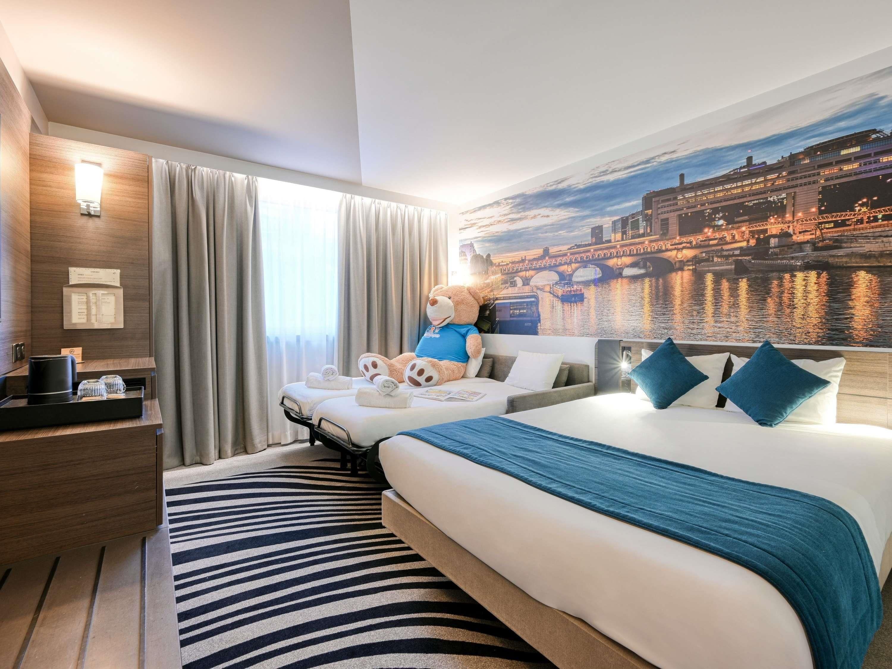 Ein Hotelzimmer mit Bett, einer Couch, einem großen Teddybären, einem Stadtbild-Wandgemälde und einem Schreibtisch mit Lampe.