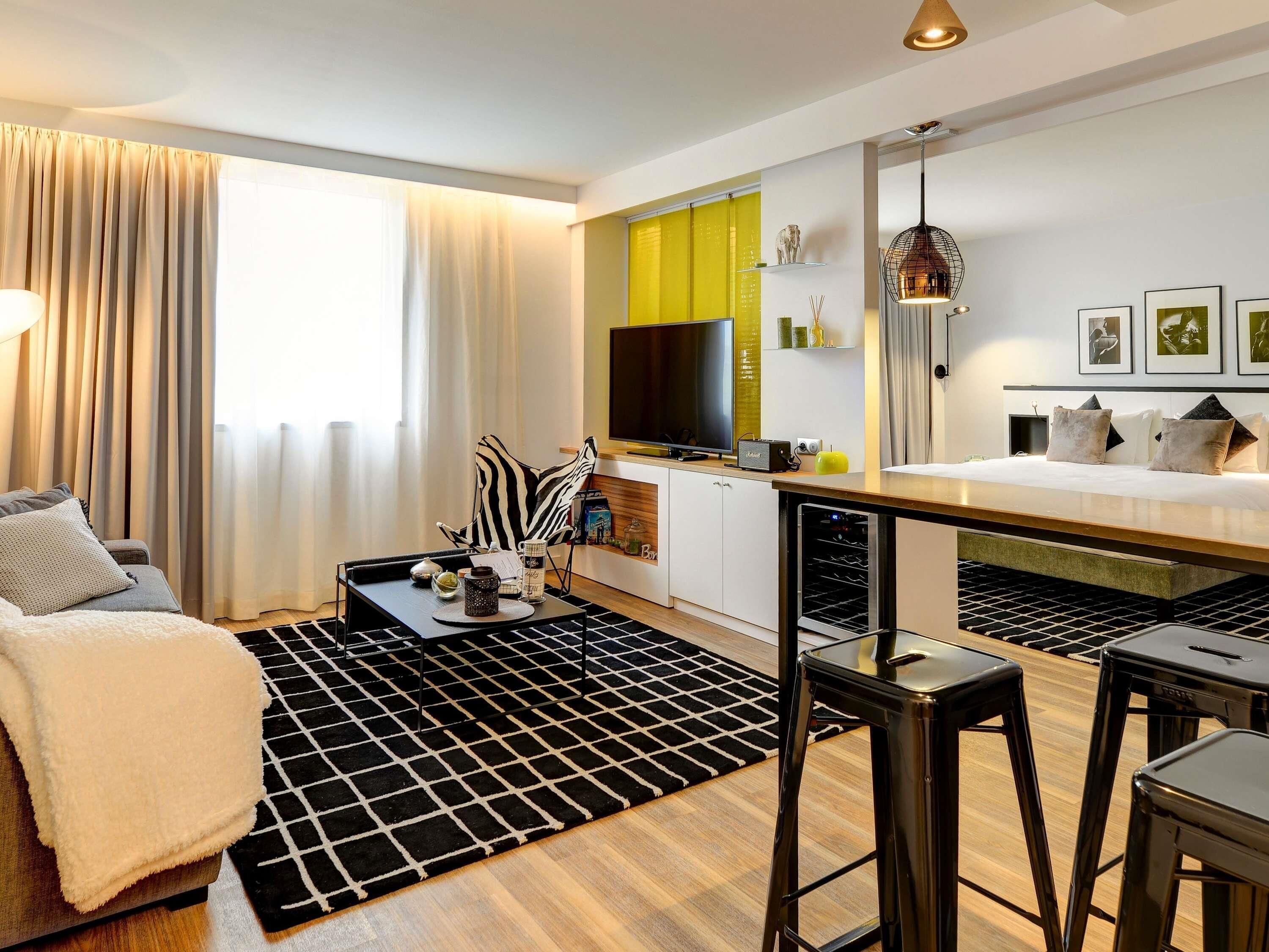 A modern living room with a sofa, coffee table, and bar stools. A flat-screen TV and a small kitchen area with a microwave are visible.