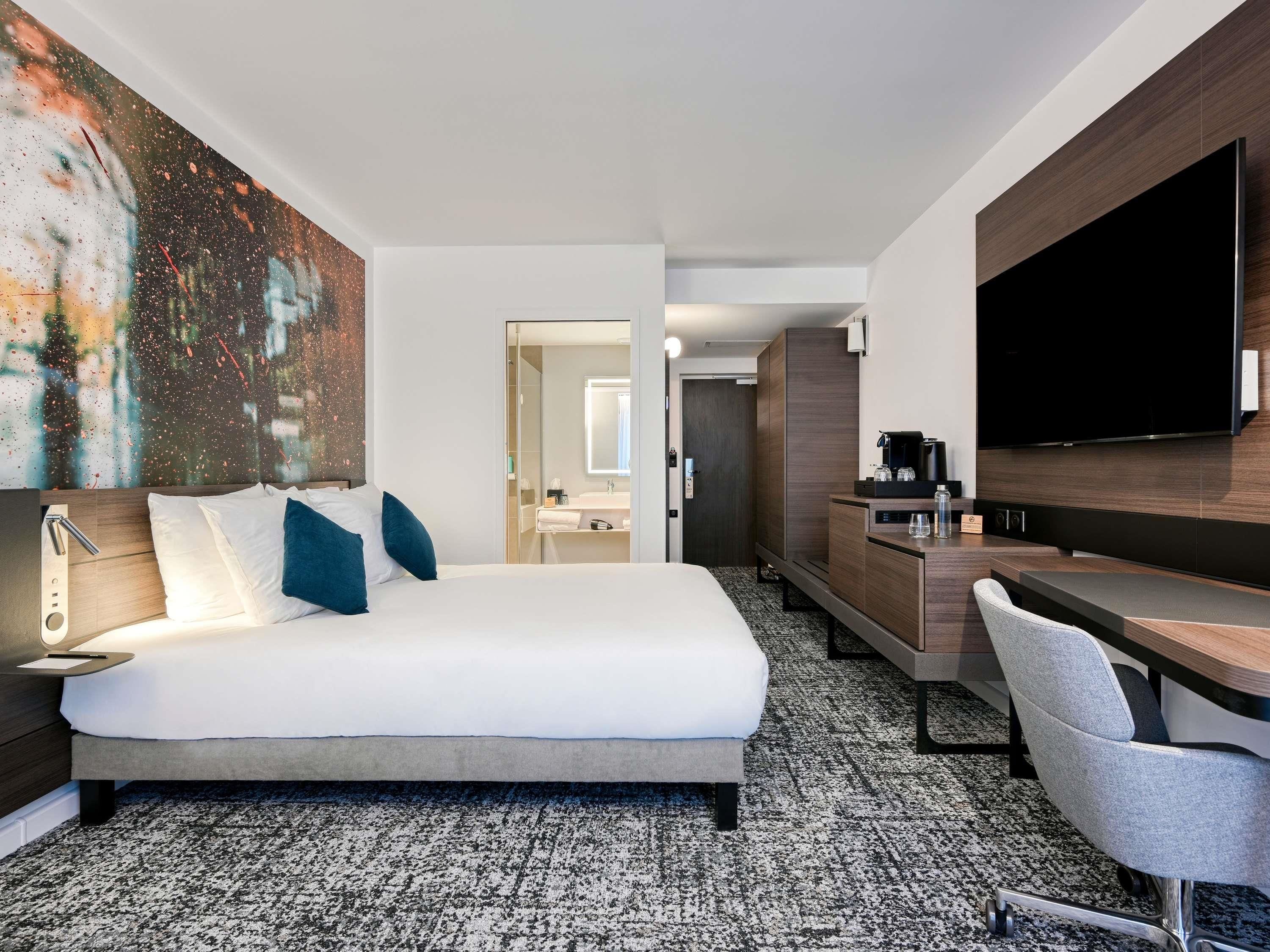 Une chambre d’hôtel avec un lit, un bureau, une télévision et une salle de bain visible à travers une porte ouverte.