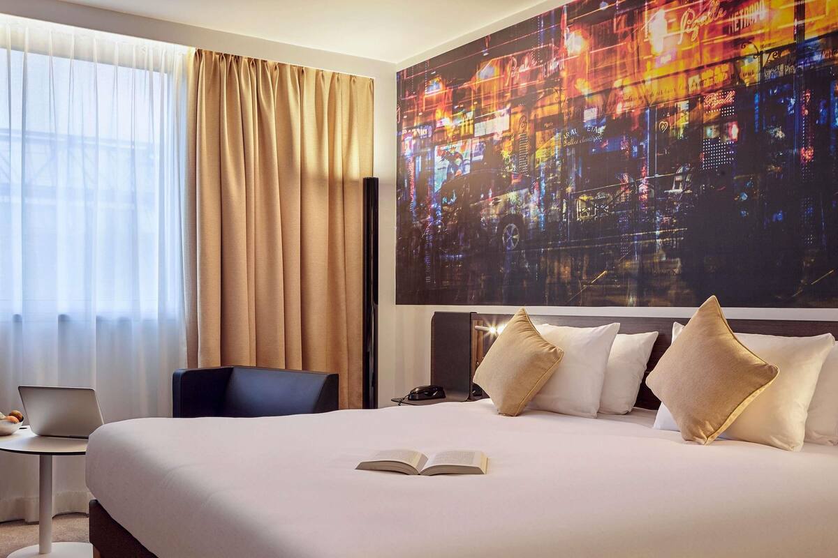 A hotel room with a large bed, a desk, a chair, and a cityscape view.