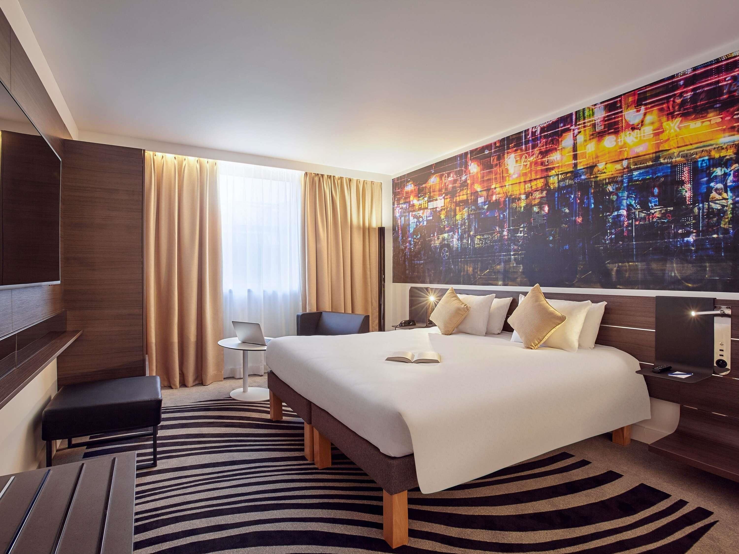 Ein Hotelzimmer mit einem großen Bett, einem Schreibtisch, einem Sessel, einem Fernseher und einem Wandbild mit Stadtansicht.