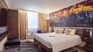 Ein Hotelzimmer mit einem großen Bett, einem Schreibtisch, einem Sessel, einem Fernseher und einem Wandbild mit Stadtansicht.