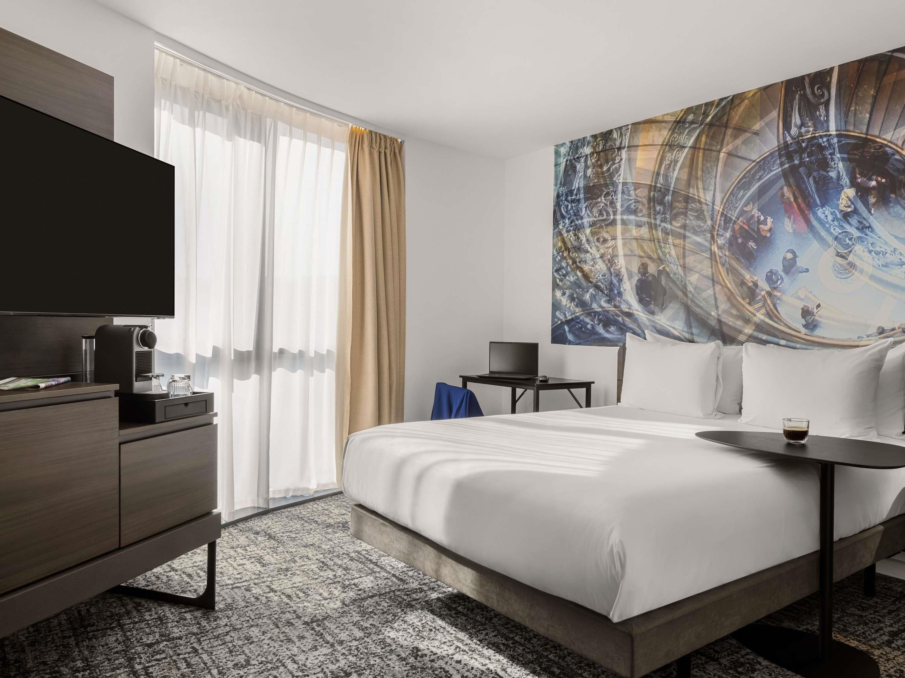 A hotel room with a bed, a desk, a television, and a large abstract painting on the wall.