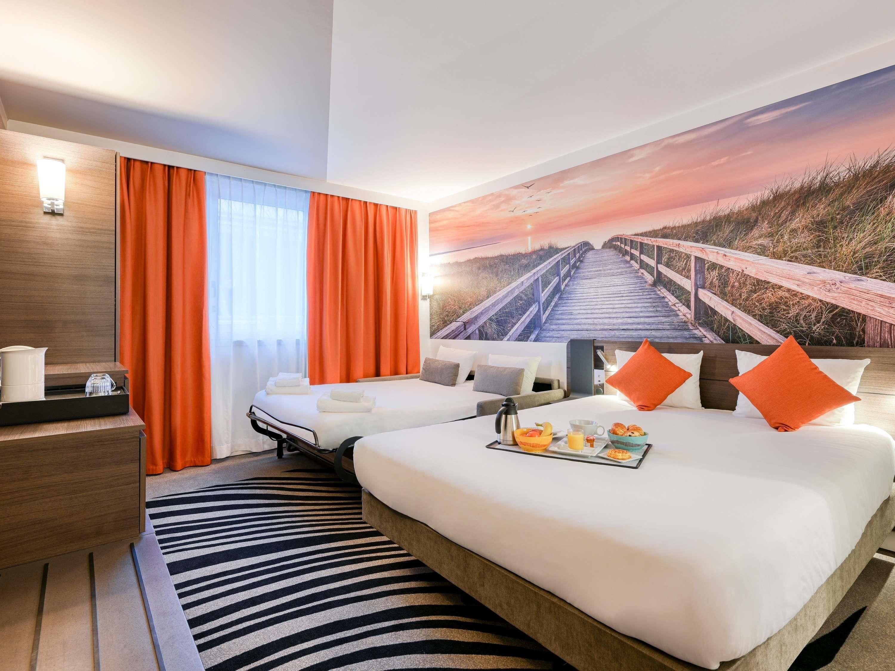 Une chambre d’hôtel avec deux lits, une table de chevet en bois, un tapis à motifs zébrés et une fresque représentant un coucher de soleil sur le mur.